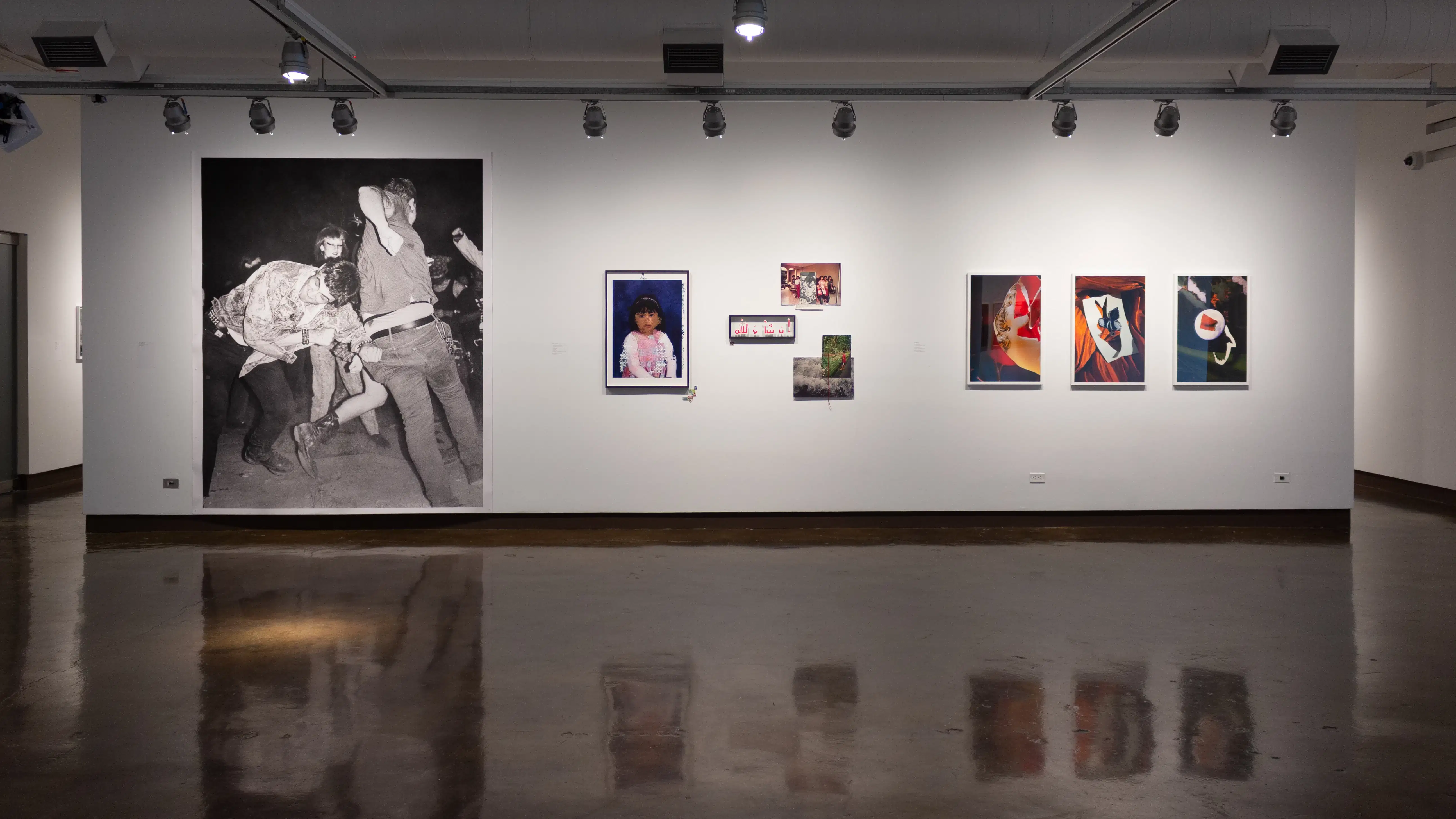 Installation image showing a wall with a large black and white photograph and 4 framed color photographs and some unframed photo installations with hallways on either side.