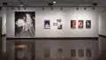 Installation image showing a wall with a large black and white photograph and 4 framed color photographs and some unframed photo installations with hallways on either side.