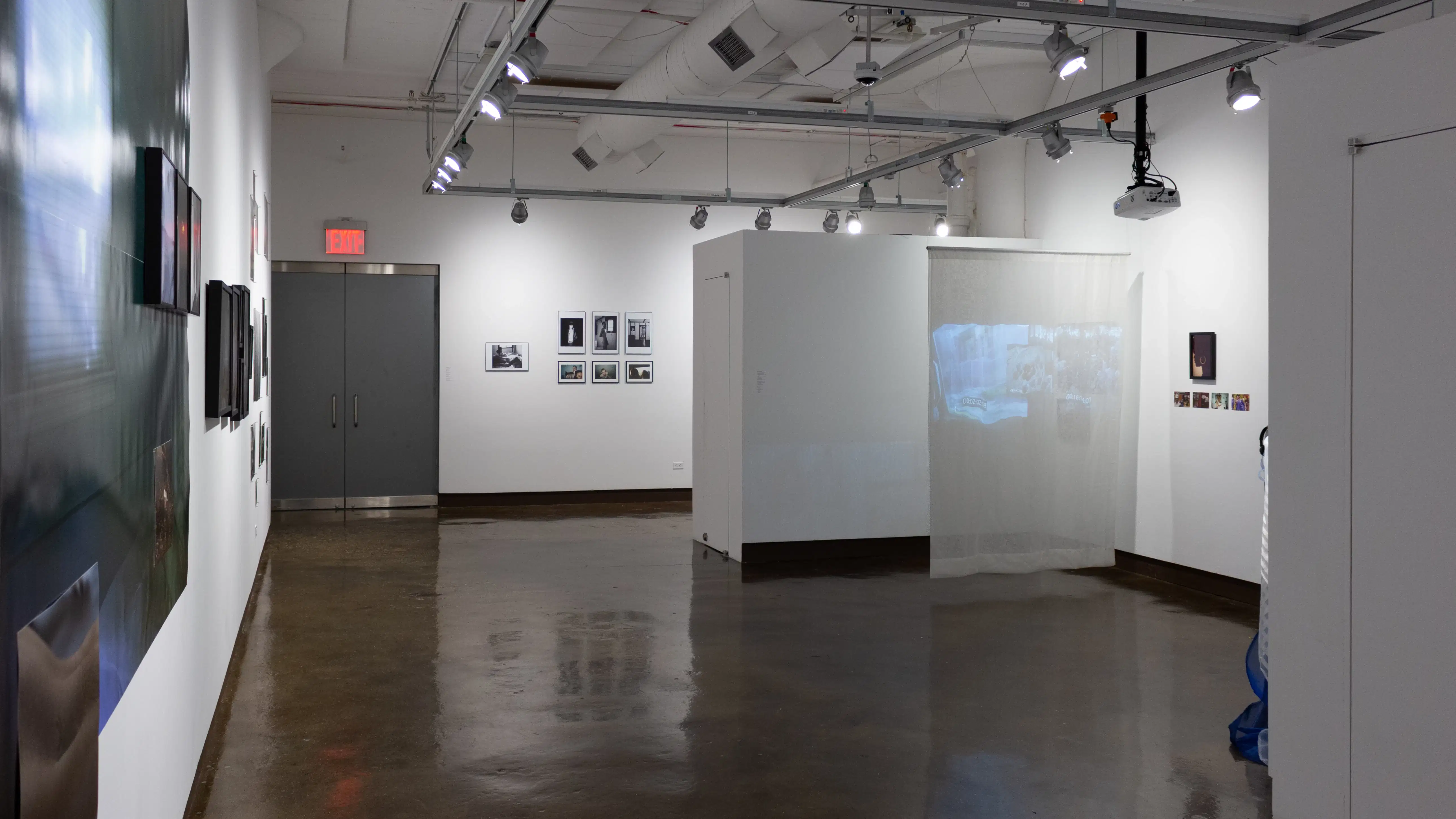 Installation image showing a projection on a hanging screen on the right, 7 framed photos on the back wall and a large framed and unframed installation on the left wall.