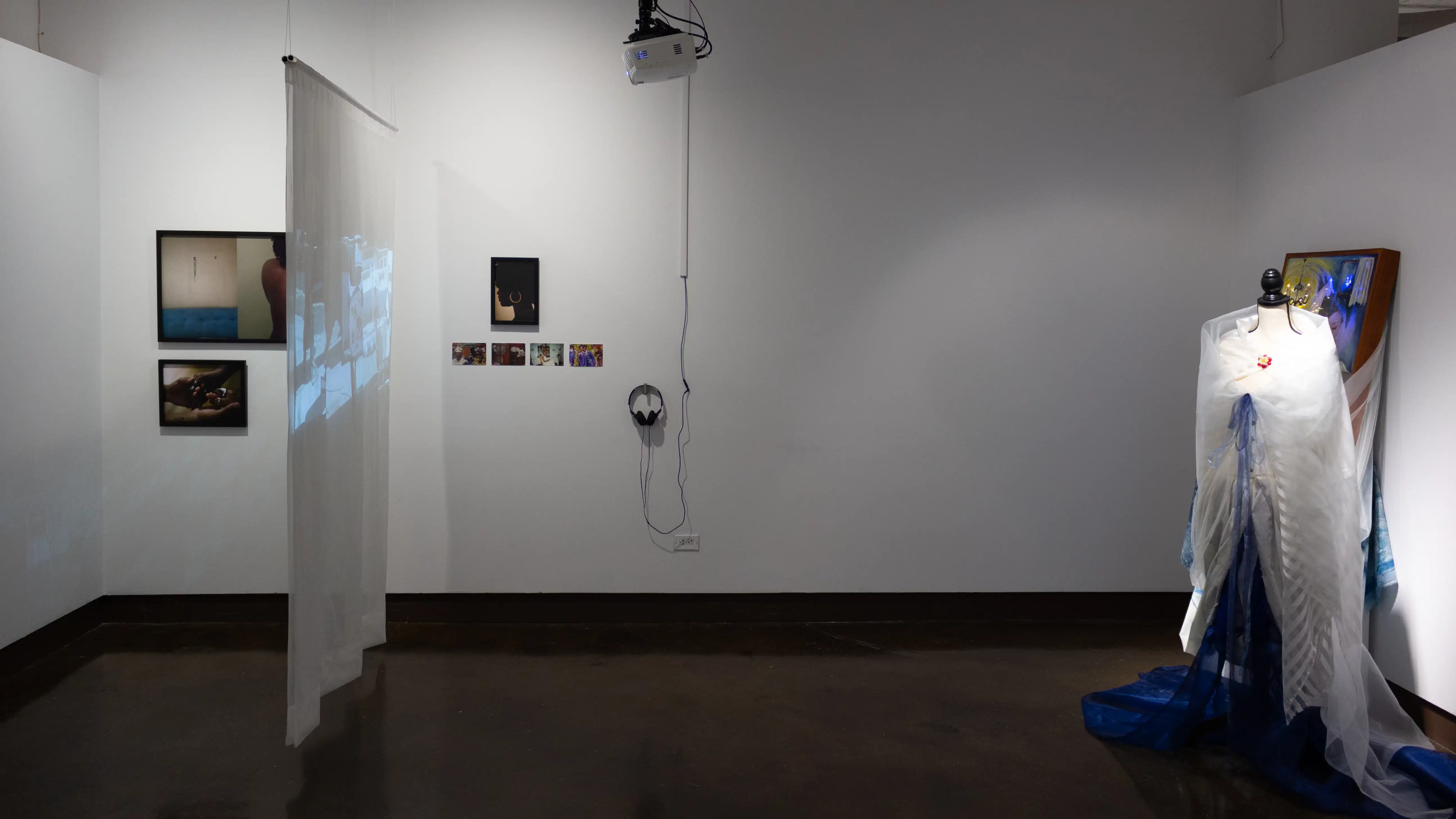 Installation image showing a projection on a hanging screen and a photo installation on the left, headphones on the wall in the middle, and a mannequin in a gown on the right.