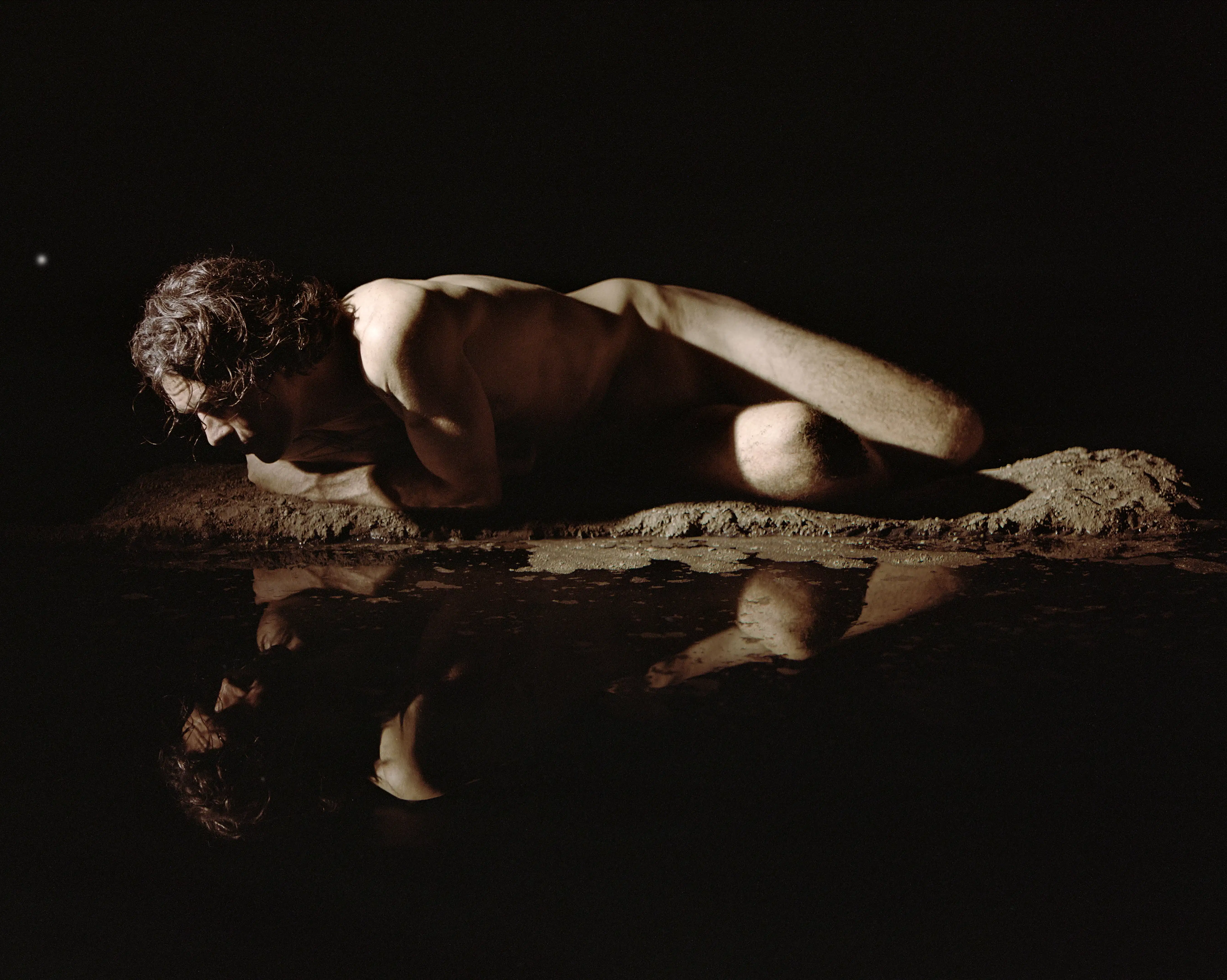 Nude man looking at reflection in water laying on rock surrounded by darkness.