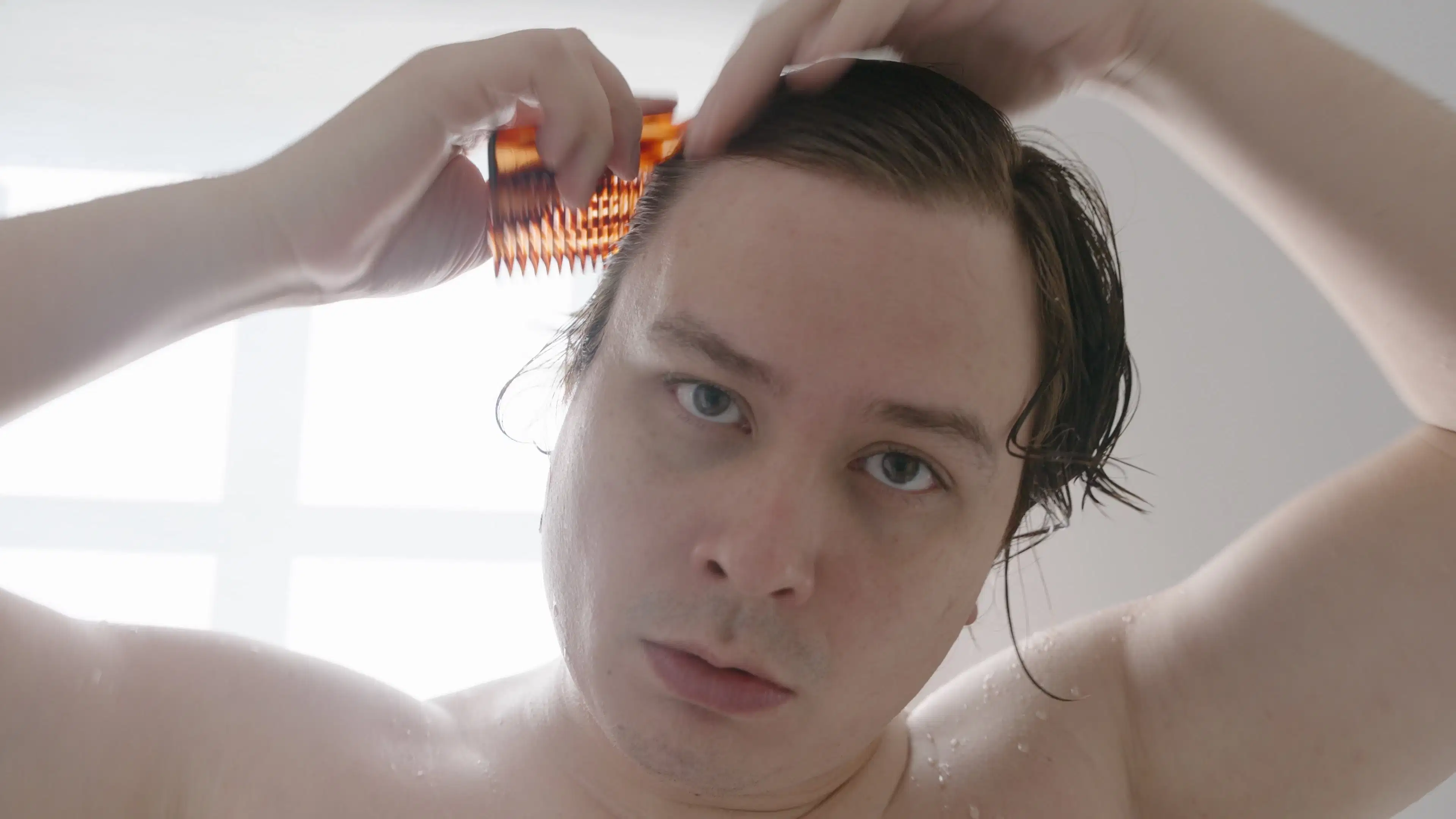 Man brushing comb through his hair, looking straight at the camera, backlit by morning light 