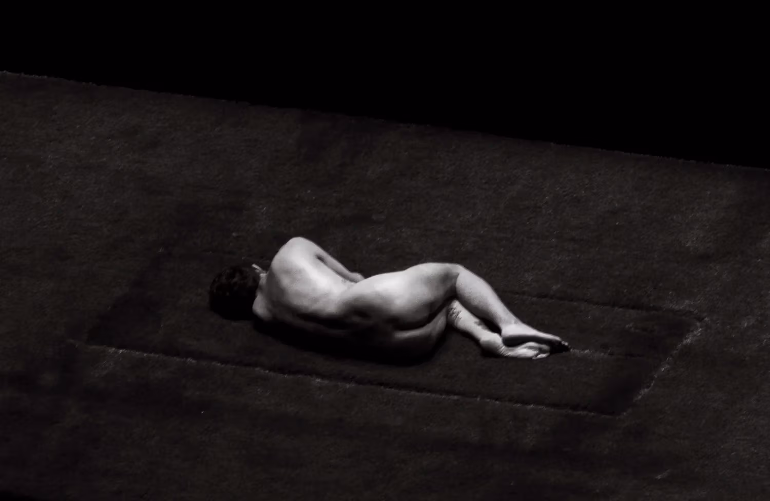 black and white image of naked man from behind in curled position, laying on grass. 