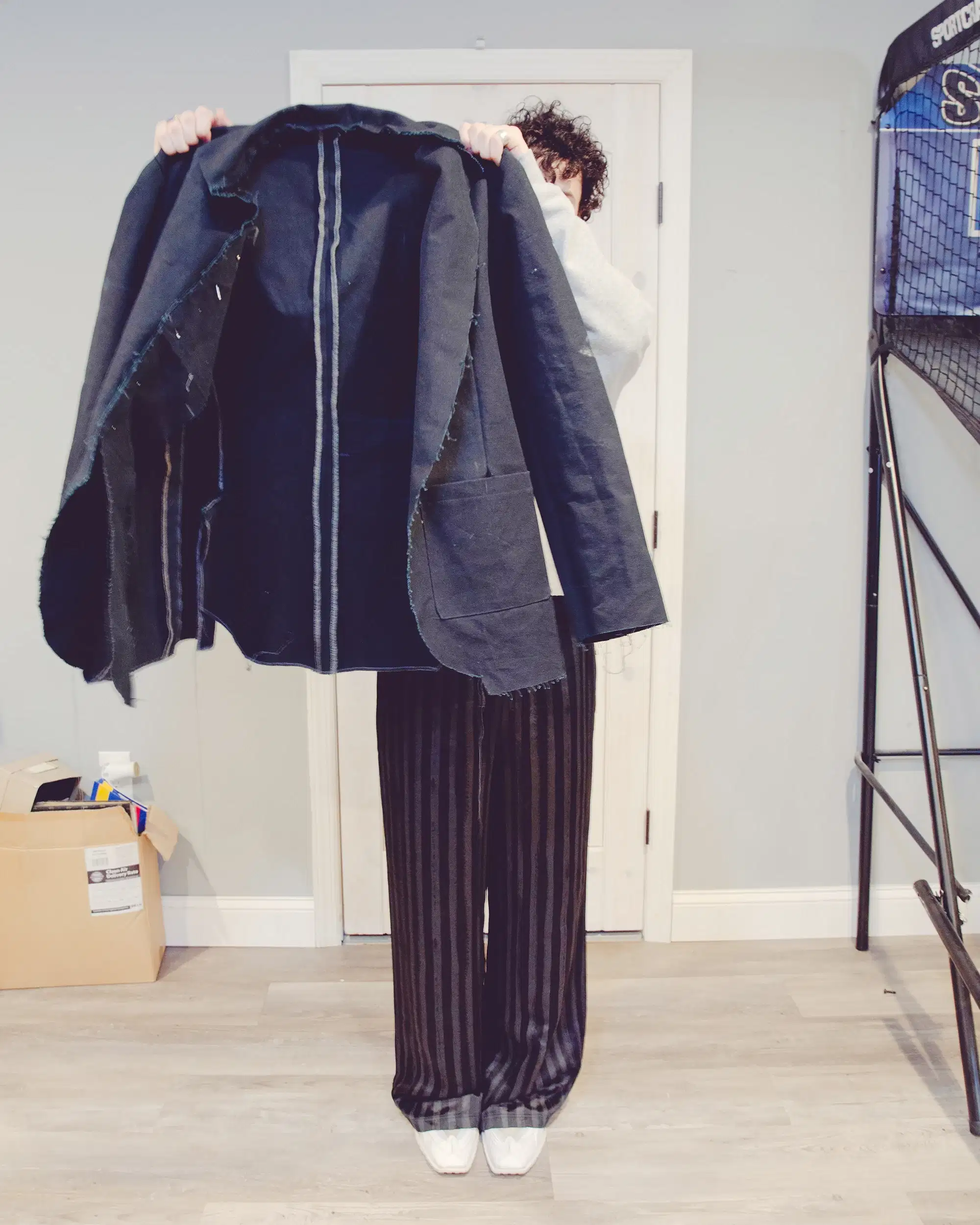 In a modern suburban basement, a young man holds up a black canvas jacket that he is almost done sewing.