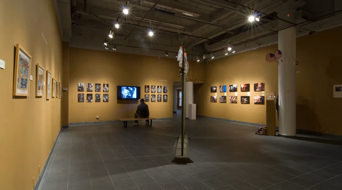 Exhibition installation view of “Testimony to War: Art from the Battlegrounds of Iraq,” February 2 - March 8, 2008, showing photographs and monitors on a wall in the background and a visitor watching the monitor from a bench.