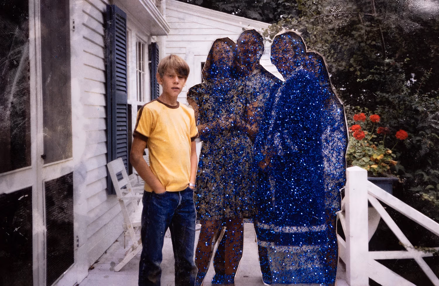 A photo featuring a young boy standing beside a cut-out of a glittering, blue silhouette of a family in front of a house with red flowers.