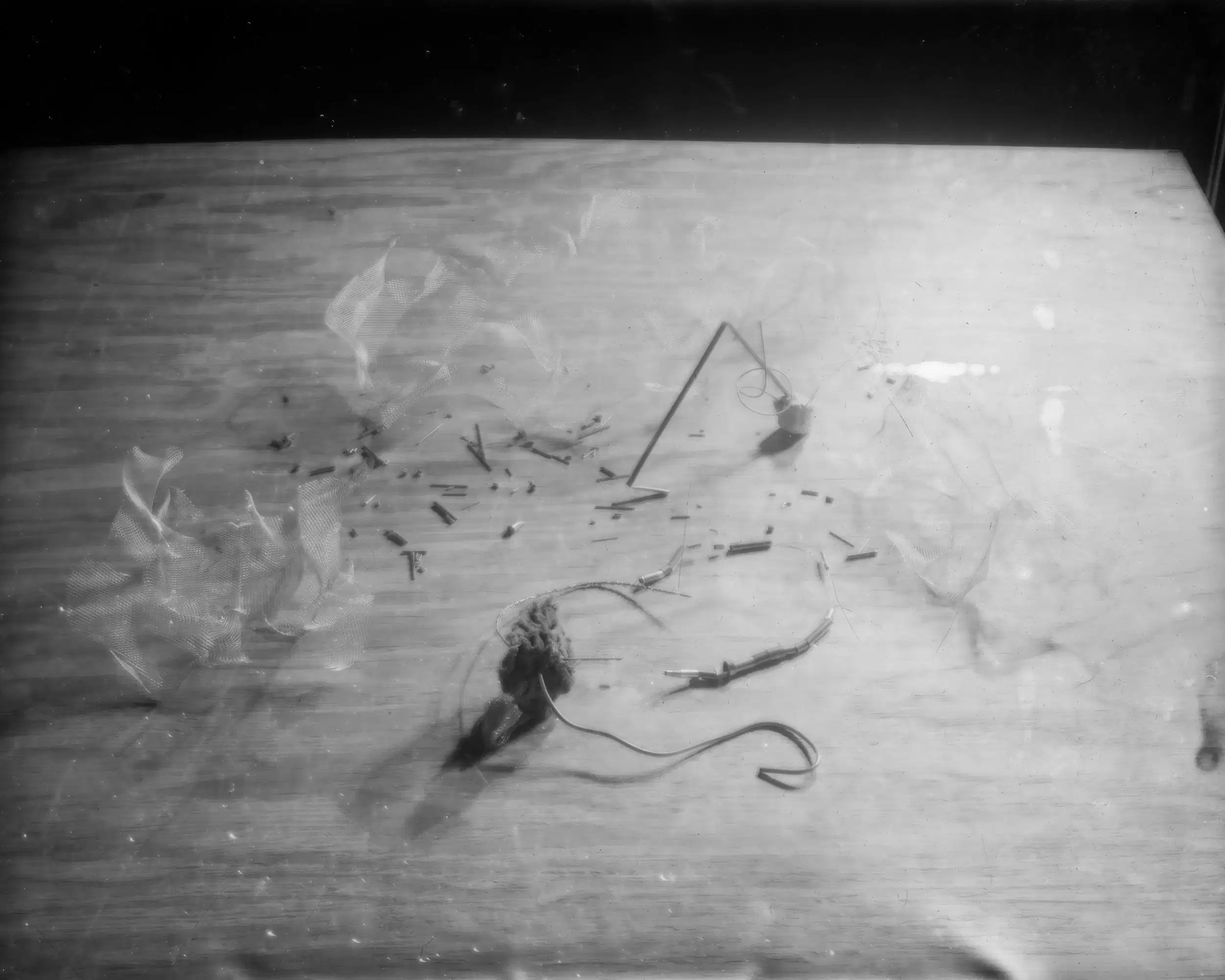 Black and white photo of scattered debris, a mesh piece, and wire on a textured wooden surface with a shadowy backdrop.