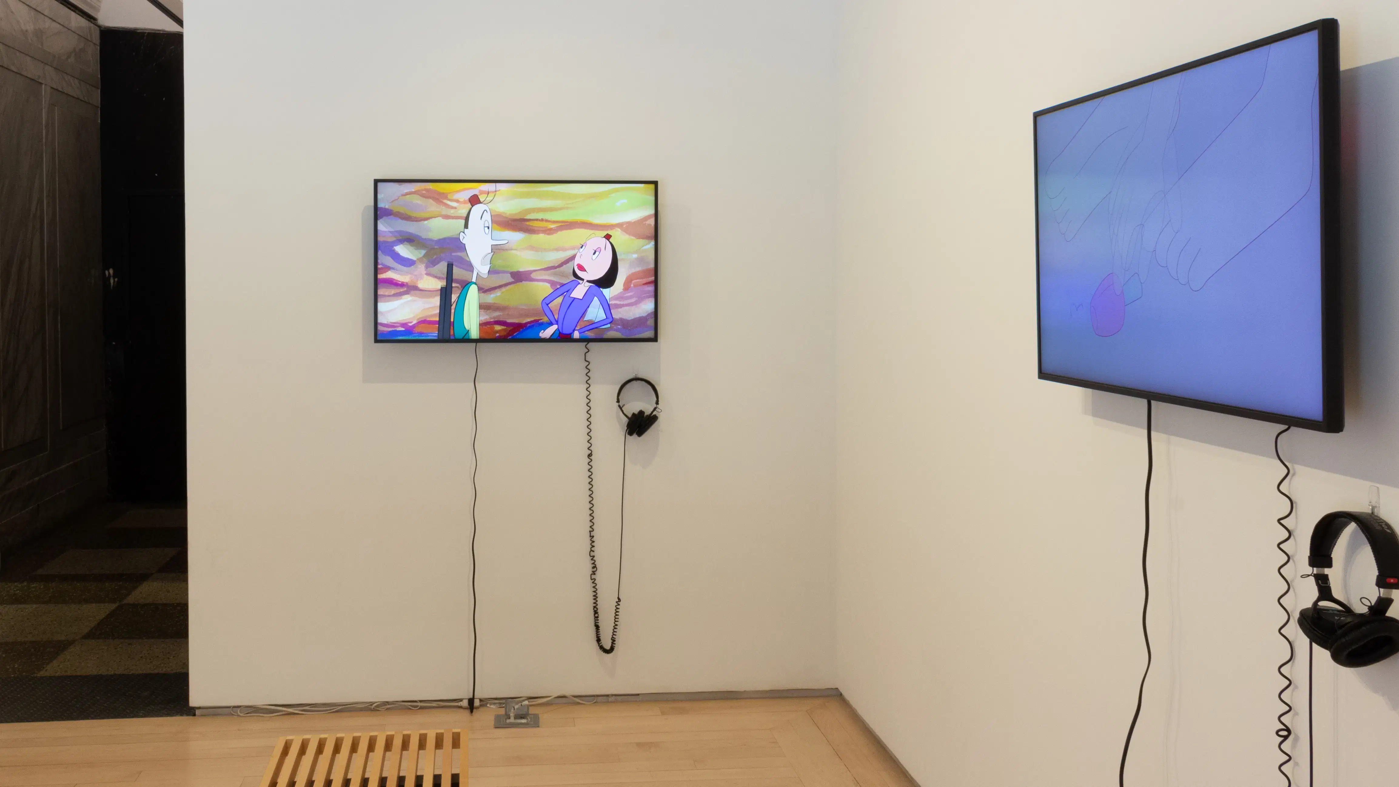Installation image showing two monitors along a wall in a small room, with a bench on the floor and a hallway leading out on the left side.