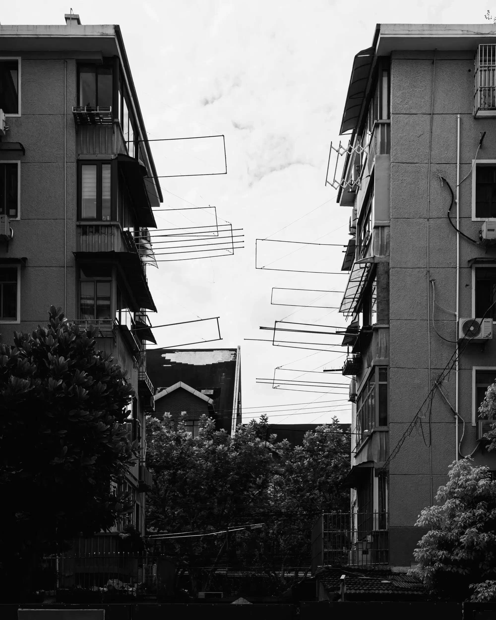 A black and white photograph capturing a narrow gap between two residential buildings. Multiple rows of empty clotheslines extend from the balconies, creating a pattern of horizontal lines against the sky. 