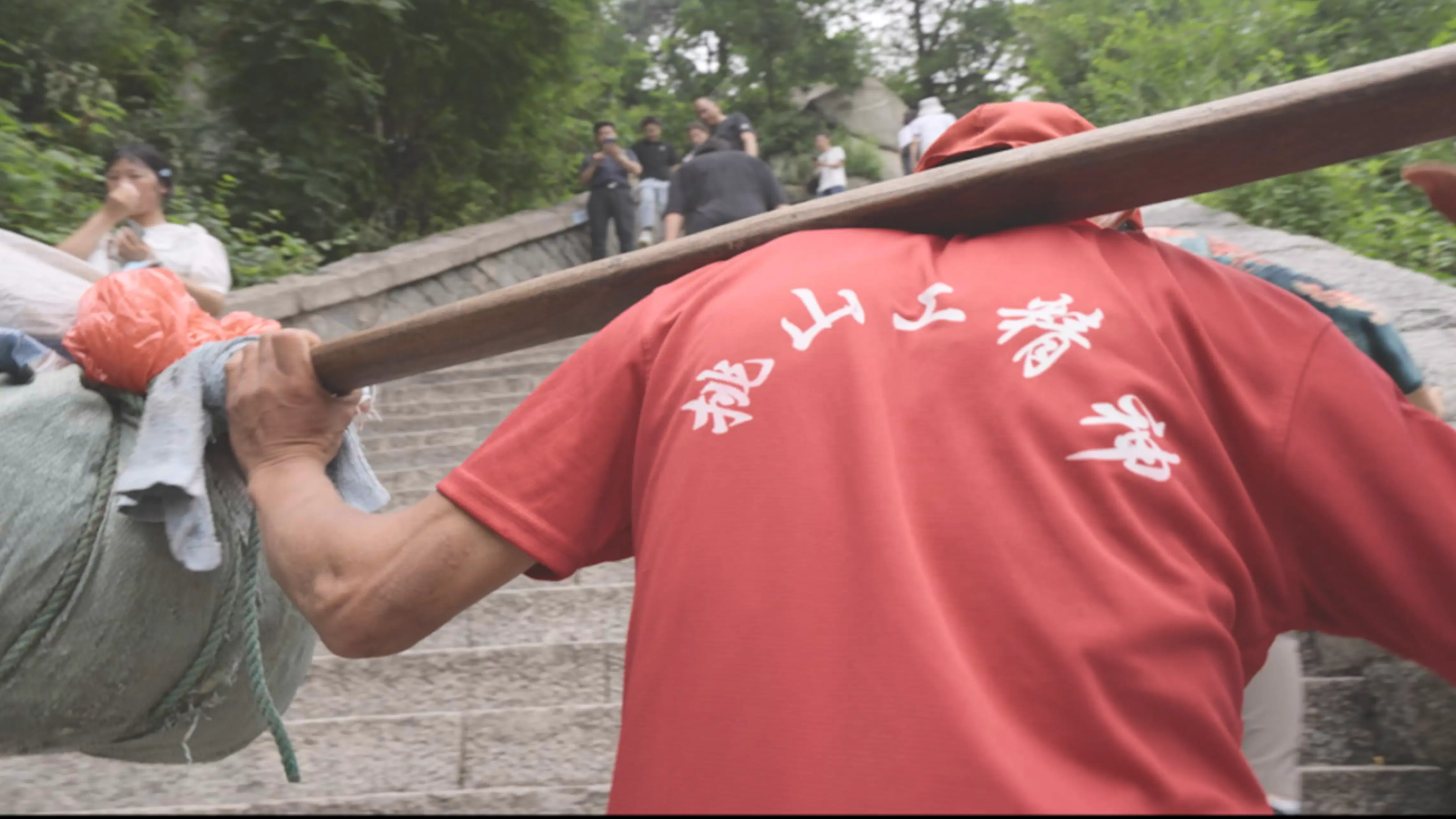 The back of a man dressed in red as he walks forward on the steps of China's Mount Tai, carrying a heavy load on a stretcher.