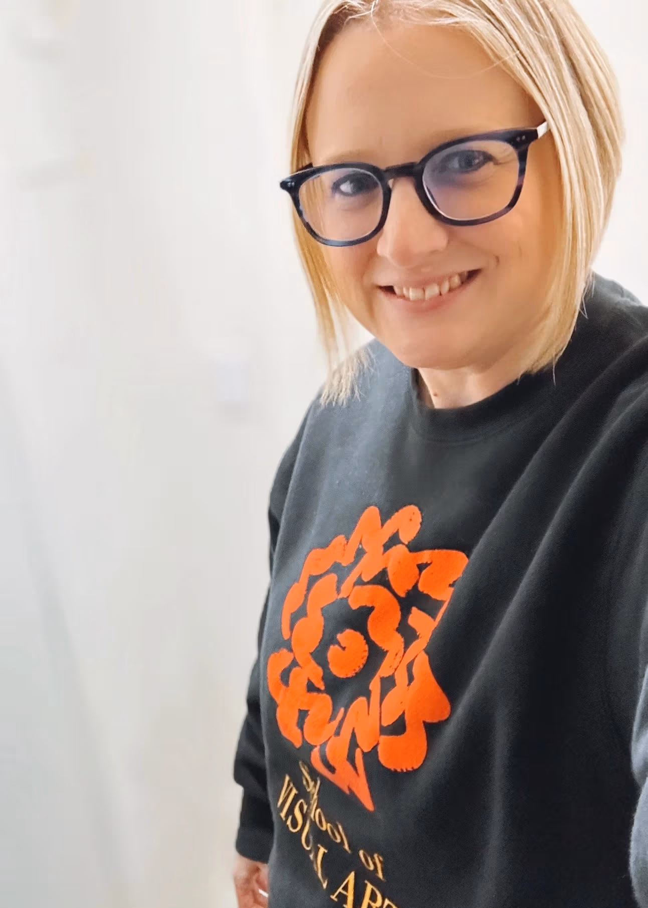 Image of Caryn, a person with chin length blonde hair and glasses who is wearing a black sweater with an orange SVA logo. 