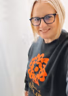 Image of Caryn, a person with chin length blonde hair and glasses who is wearing a black sweater with an orange SVA logo. 