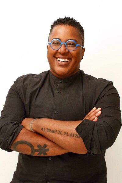 Faculty Sloan Leo smiling and standing against a white background with arms crossed wearing a black shirt and blue rimmed glasses.