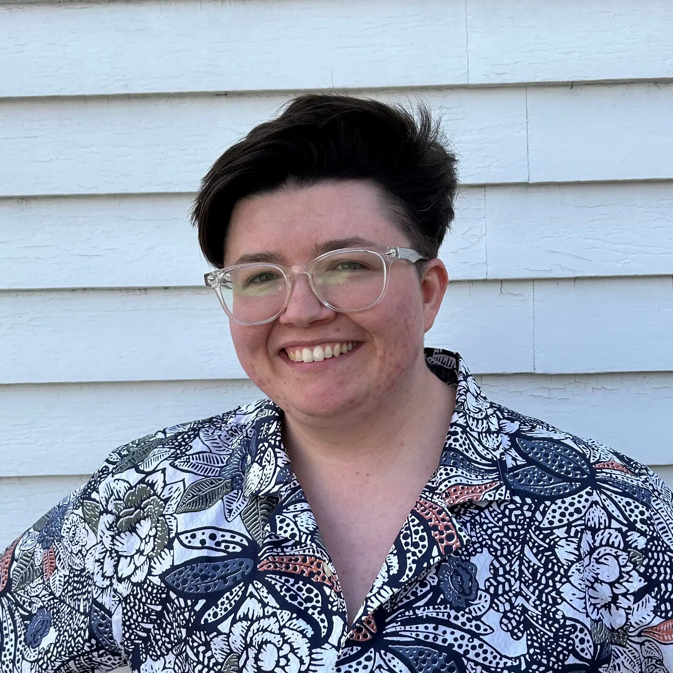 An image of Kara, a non-binary person with short brown hair and glasses. Kara is wearing a button down shirt with large white, blue, or pinkish flowers. 