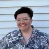 An image of Kara, a non-binary person with short brown hair and glasses. Kara is wearing a button down shirt with large white, blue, or pinkish flowers. 