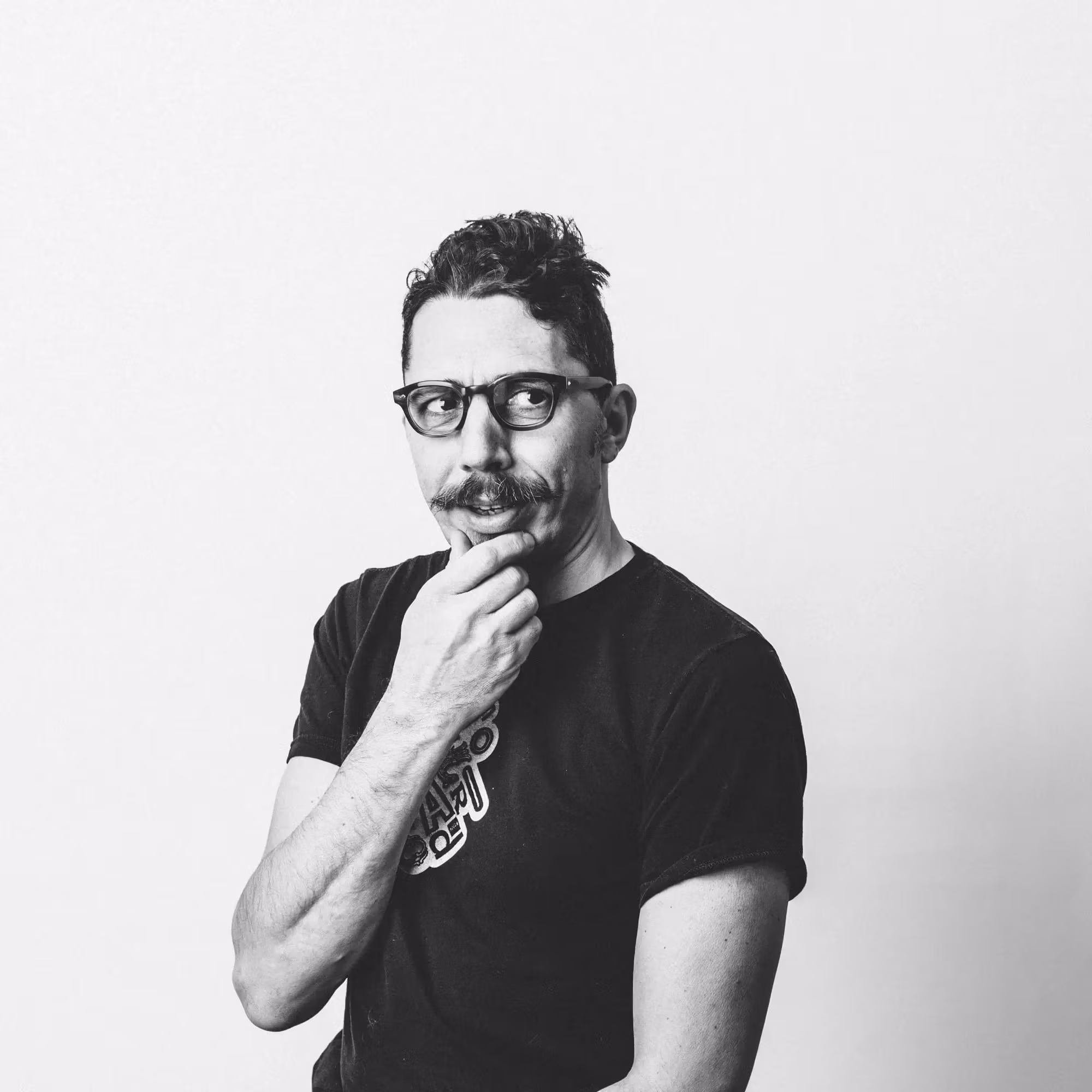 A man with a mustache and dark rimmed glasses looks off camera and hold his chin in his hand. The image is black and white.