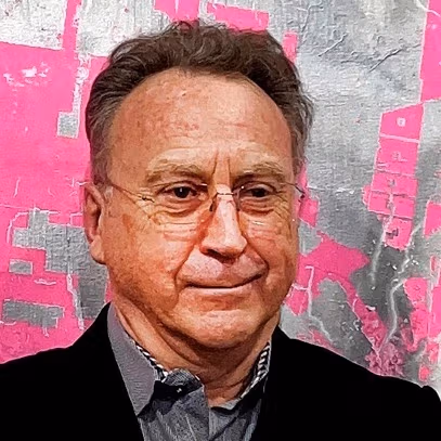 An older white man with brown hair and glasses smiling looking away from the camera. 
