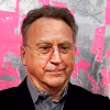 An older white man with brown hair and glasses smiling looking away from the camera. 
