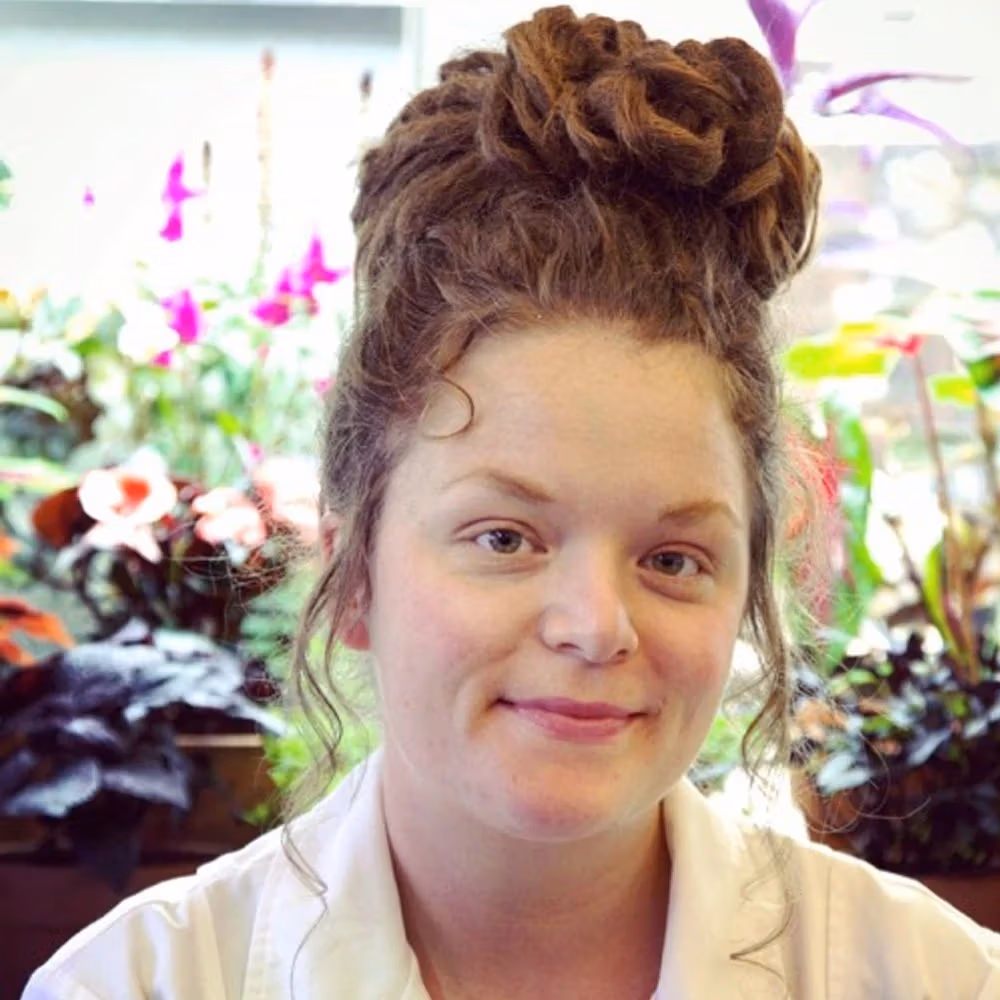 A white woman with red hair smiles at the camera and stands in front of a terrarium filled with plants.