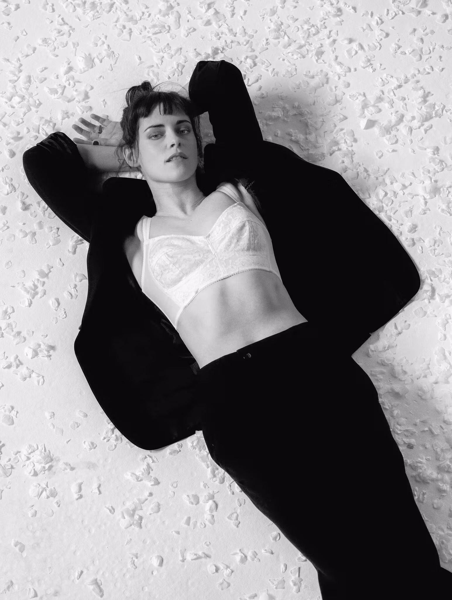 A person lies on a textured surface, wearing a dark jacket and pants over a light-colored bralette, with arms resting above their head.