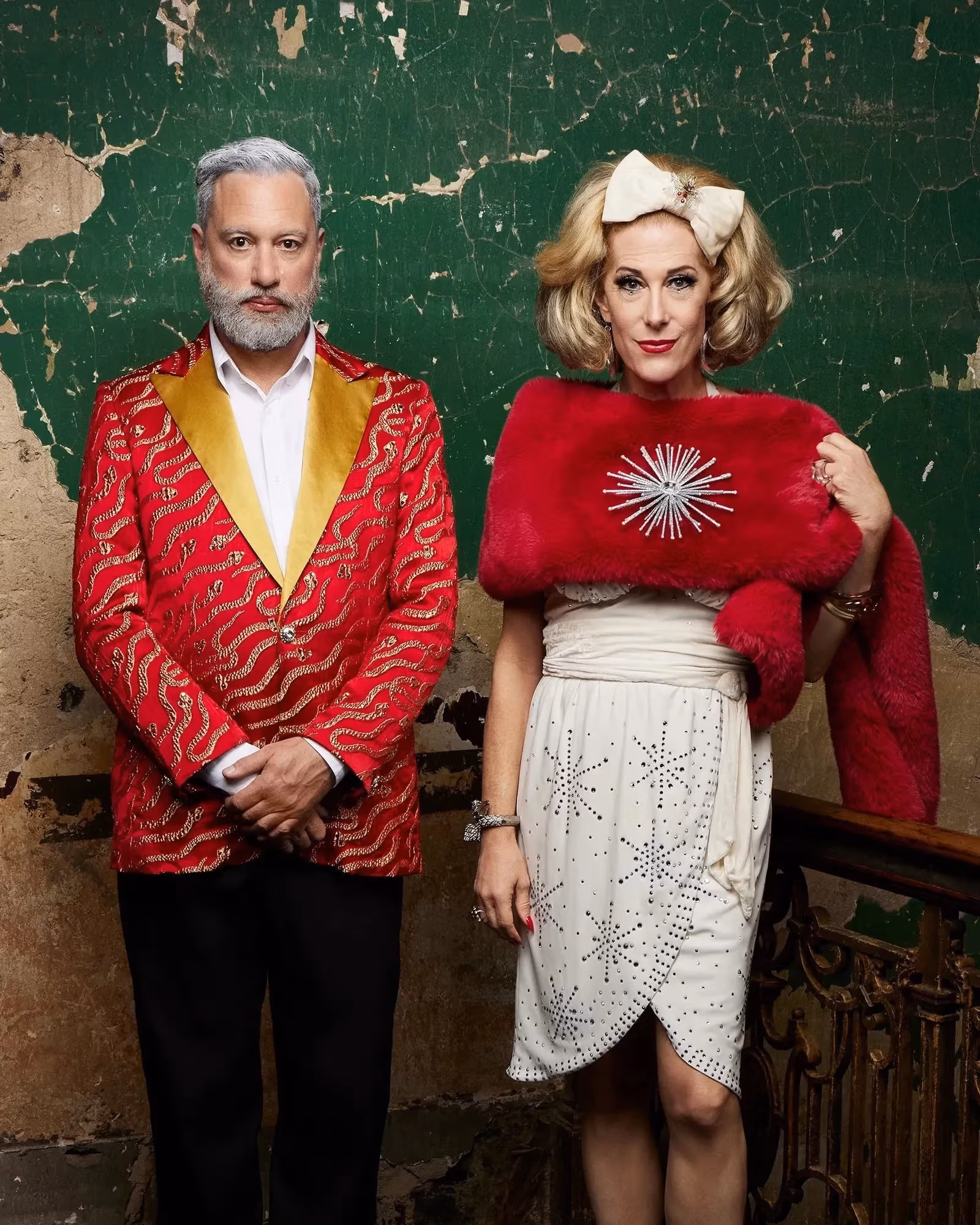 Image of a cis queer gendered man in a red patterned suit with a yellow collar and a them/ they drag performer in a white dress and red fur stole standing in front of a distressed old stucco green wall.Image of a cis queer gendered man in a red patterned suit with a yellow collar and a them/ they drag performer in a white dress and red fur stole standing in front of a distressed old stucco green wall.v
