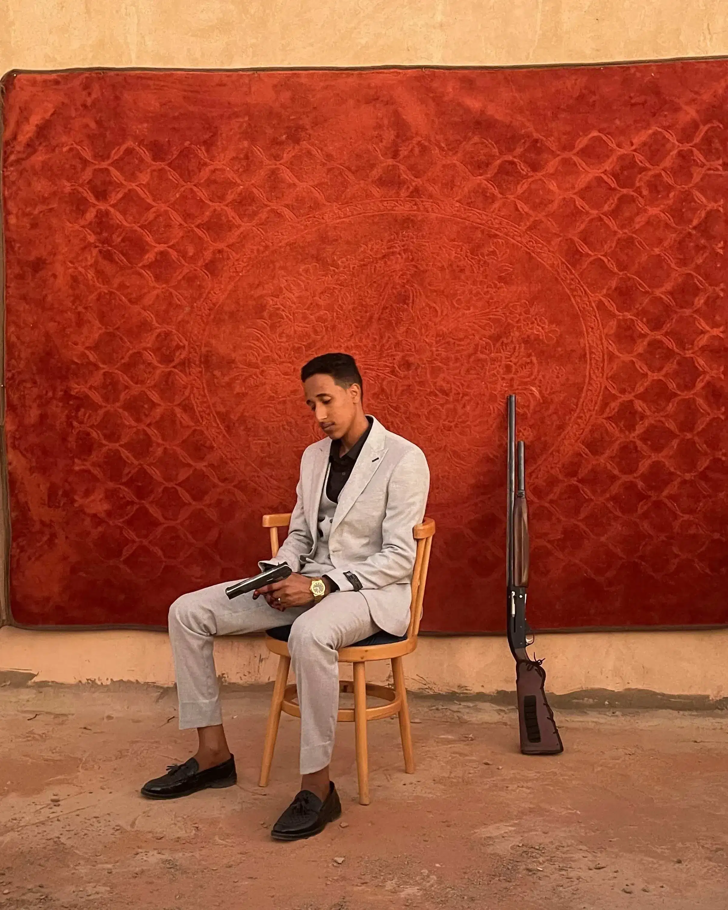 A man in a light suit sits on a wooden chair reading, in front of a red patterned rug on a sand-colored wall. A rifle leans beside him, creating a contemplative and serene tone.