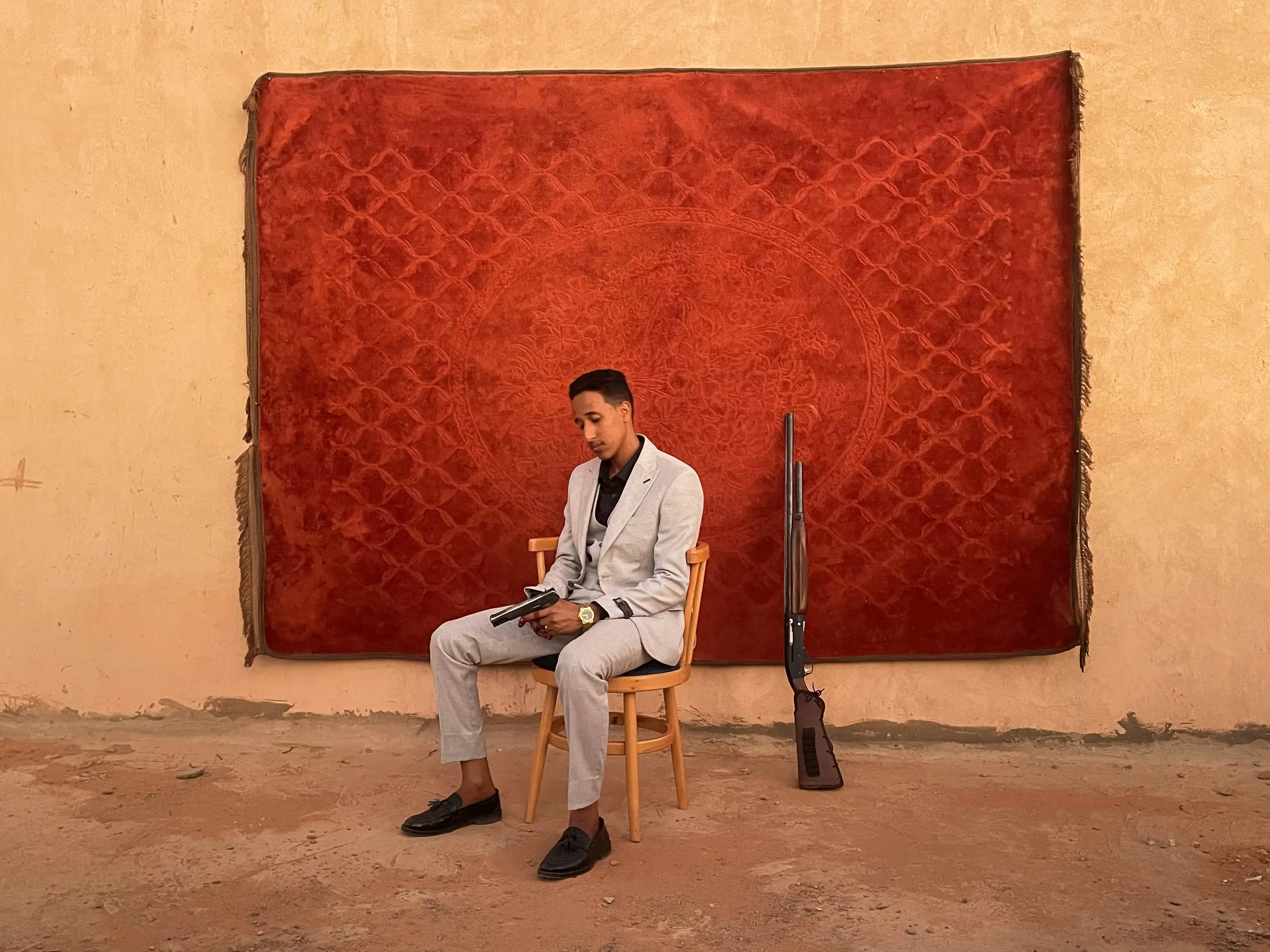 A man in a light suit sits on a wooden chair reading, in front of a red patterned rug on a sand-colored wall. A rifle leans beside him, creating a contemplative and serene tone.