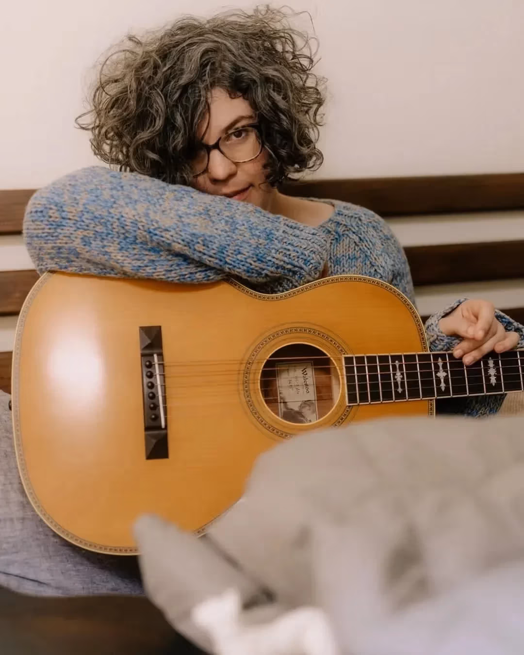 A person with curly hair and glasses leans on an acoustic guitar, wearing a blue sweater. The mood is relaxed and introspective.