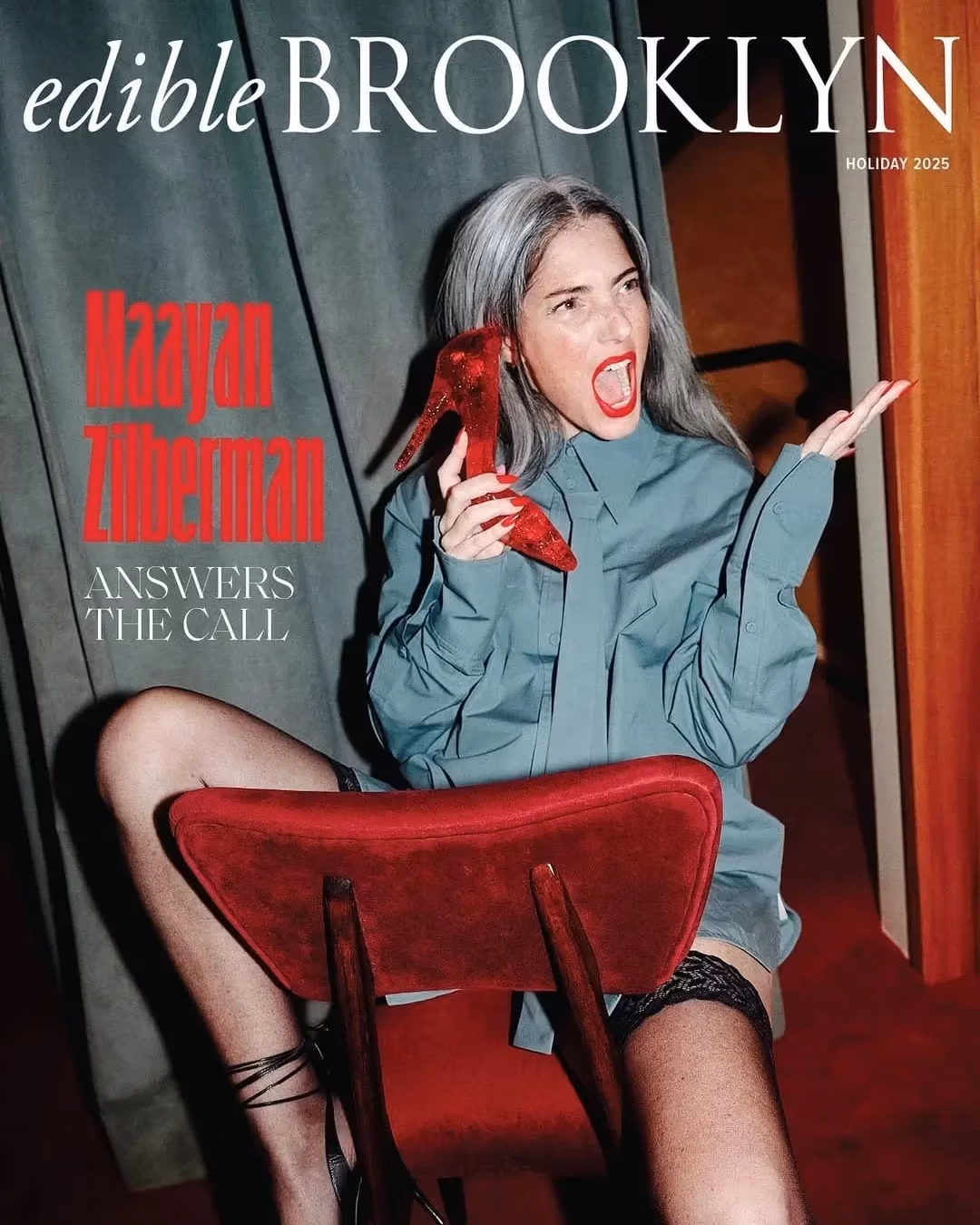 A person sitting backward on a red chair, holding a red high heel to her ear, laughing. Wears a blue shirt and black stockings, with gray hair and red lipstick.