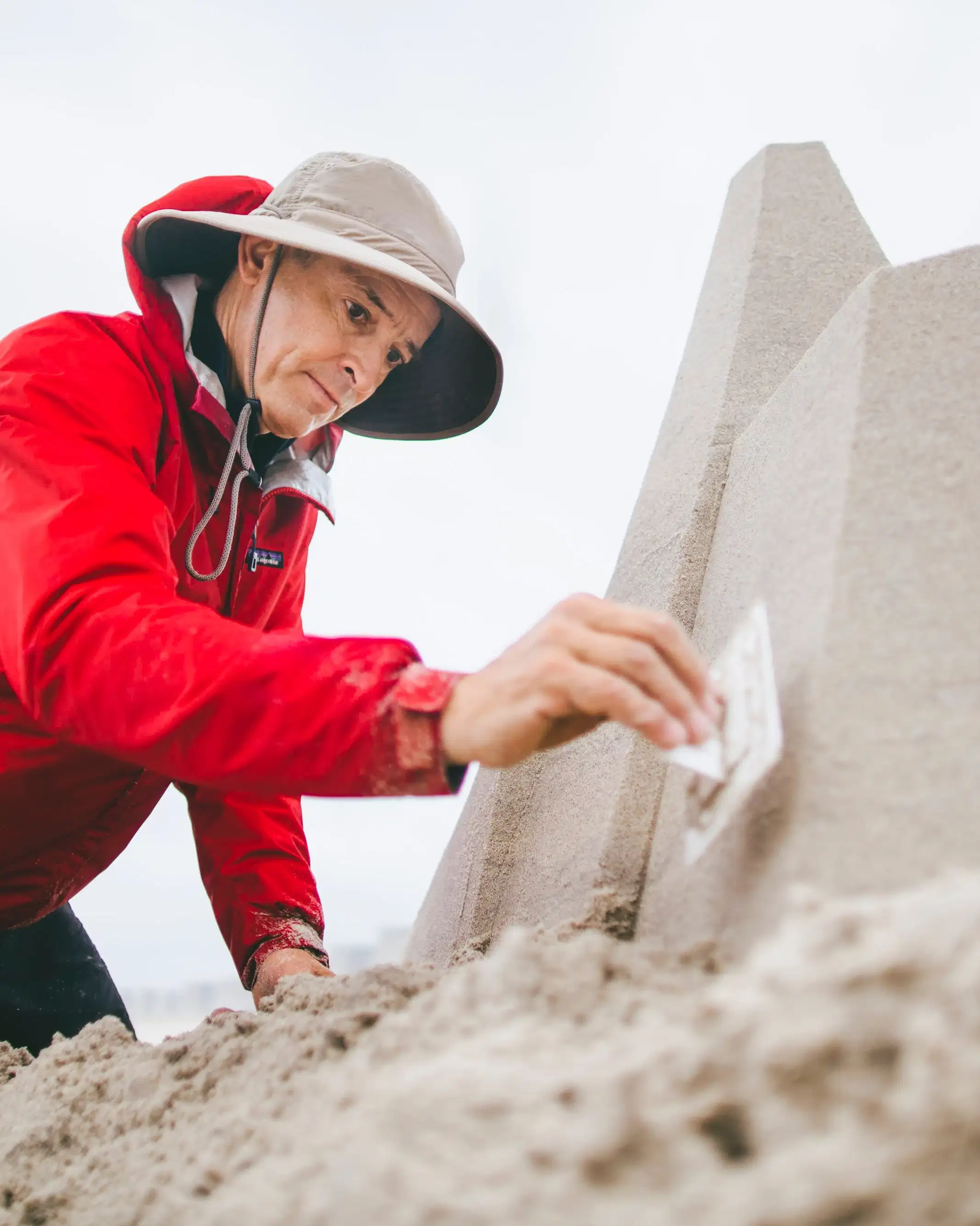 A man in a raincoat, hat, and shorts, kneels in the sand on a beach and works on a pyramidical sandcastle. He is tamping the sand down on one wall of the structure using a rectangular piece of plastic. 