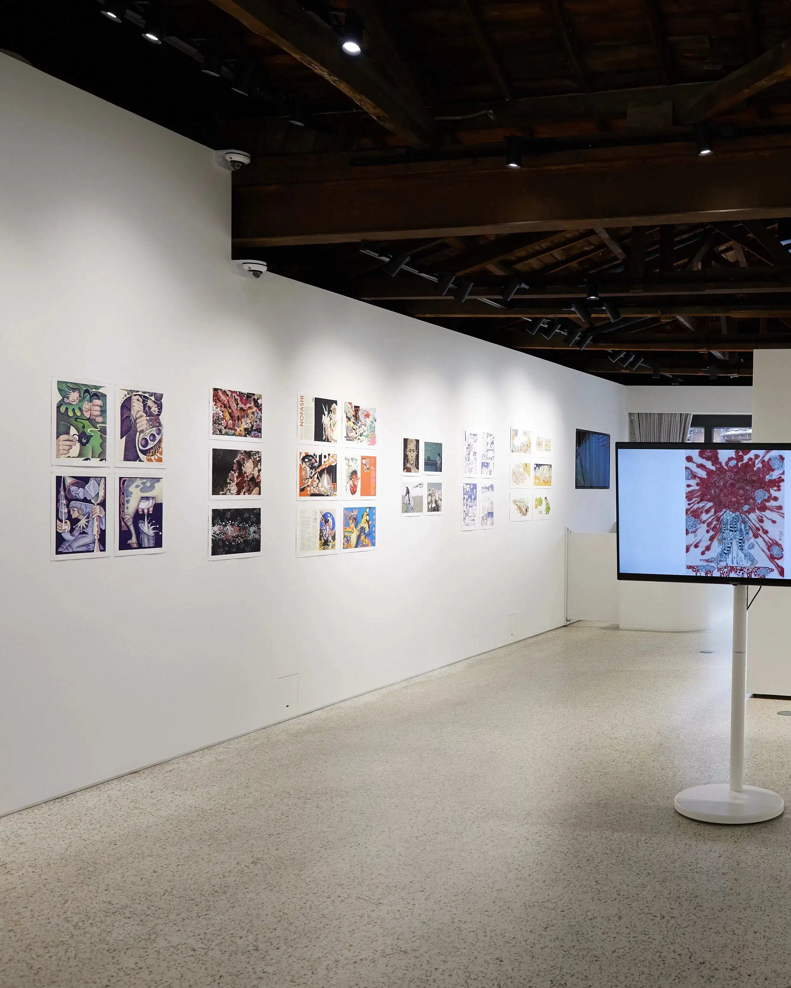 Gallery installation displaying colorful comic and illustration-style artworks arranged linearly along both walls. A large monitor on a white pedestal at the center shows a figure with red abstract patterns. The space features an open ceiling with exposed dark wooden beams, track lighting, and natural light filtering through glass doors at the far end.