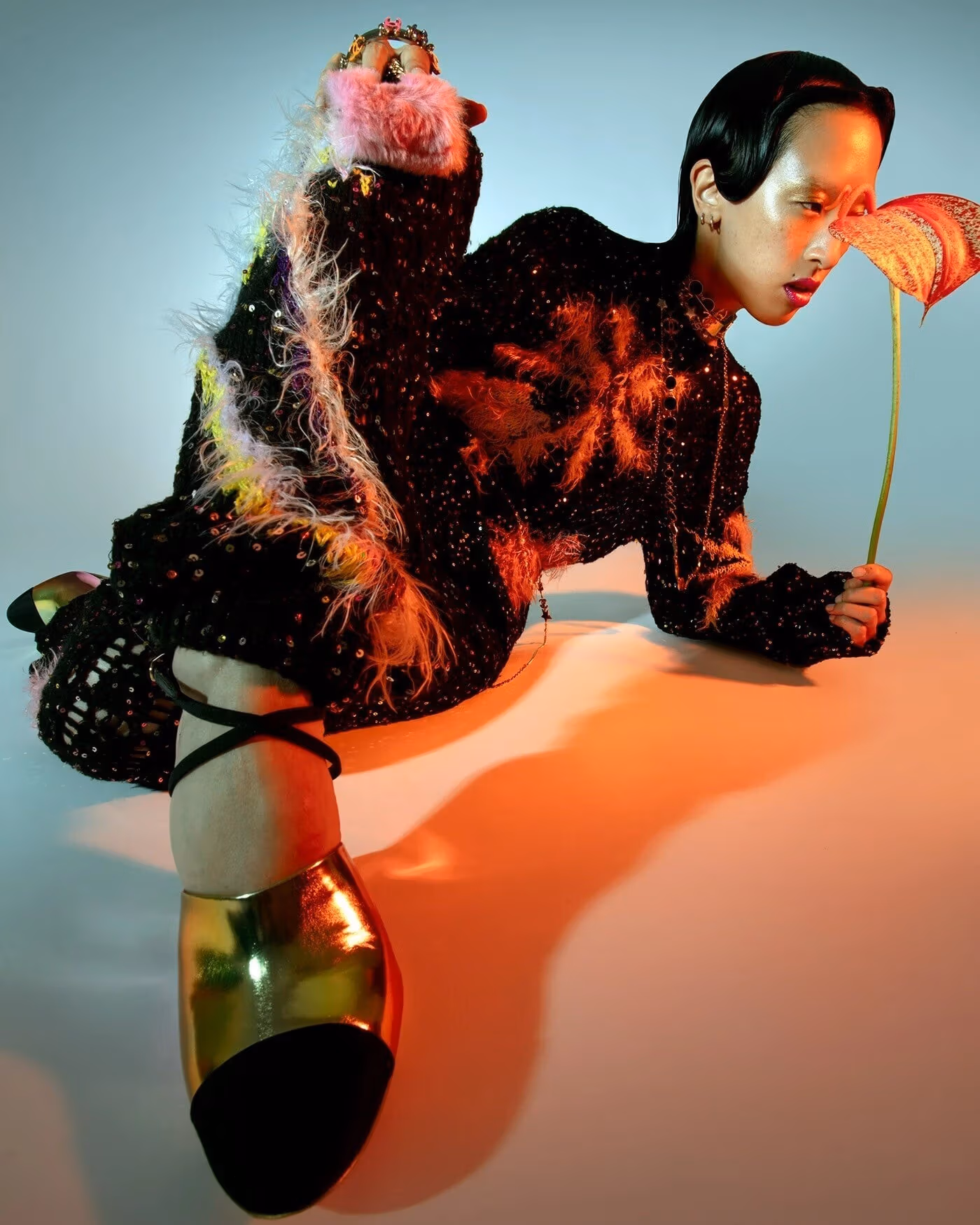 Portrait of a person sitting on the floor, posing with their right foot close to the camera and body leaning back. They hold a tropical flower in their left hand, leaning in as if to smell it. 