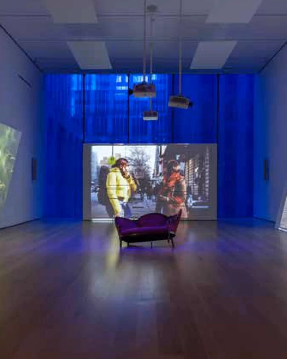 Picture of a museum installation with a couch in the middle of a room, surrounded by three big projected screens. One shows a close up of some grass, the other shows two women talking on the street, and the third one shows a park with a lake. 