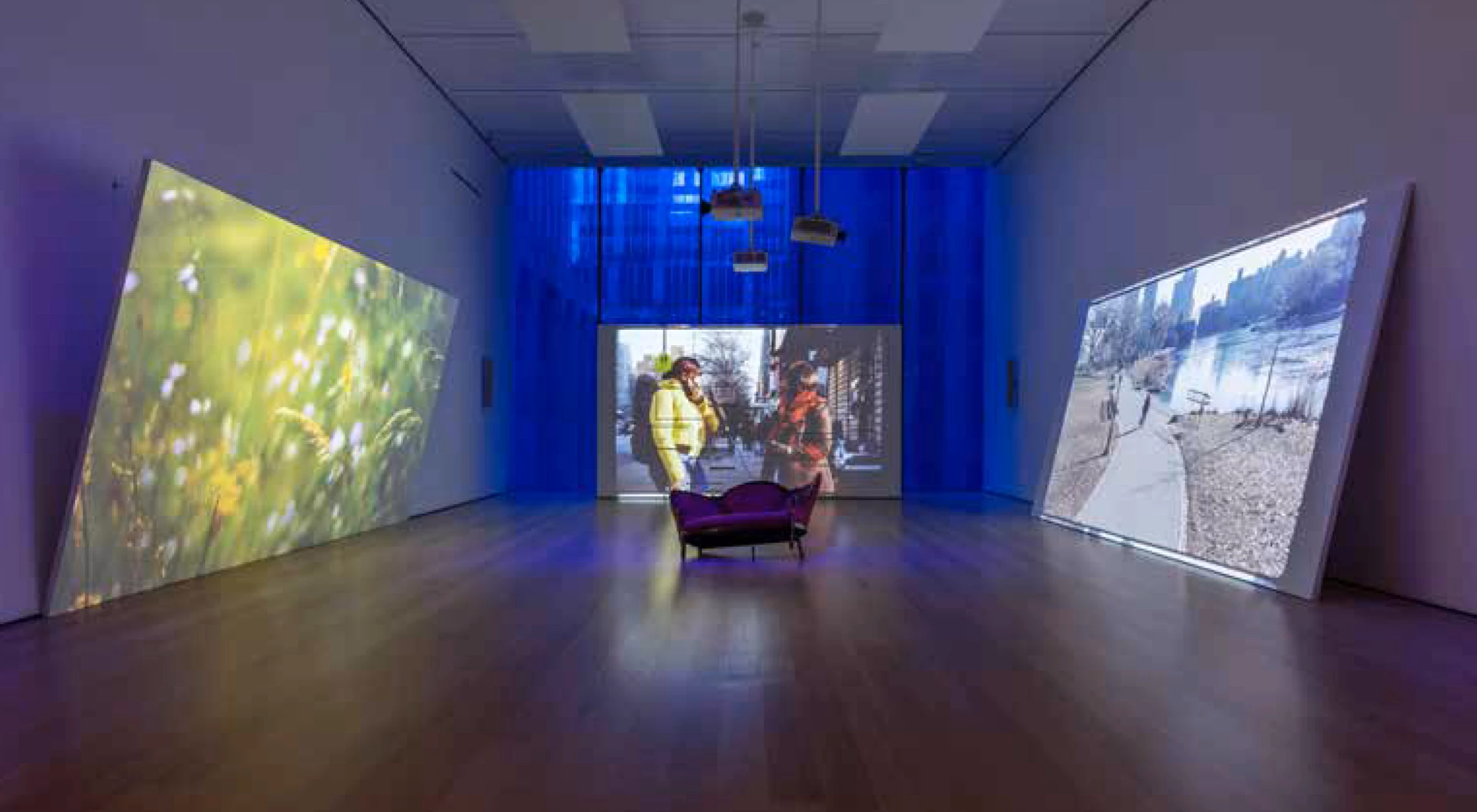 Picture of a museum installation with a couch in the middle of a room, surrounded by three big projected screens. One shows a close up of some grass, the other shows two women talking on the street, and the third one shows a park with a lake. 