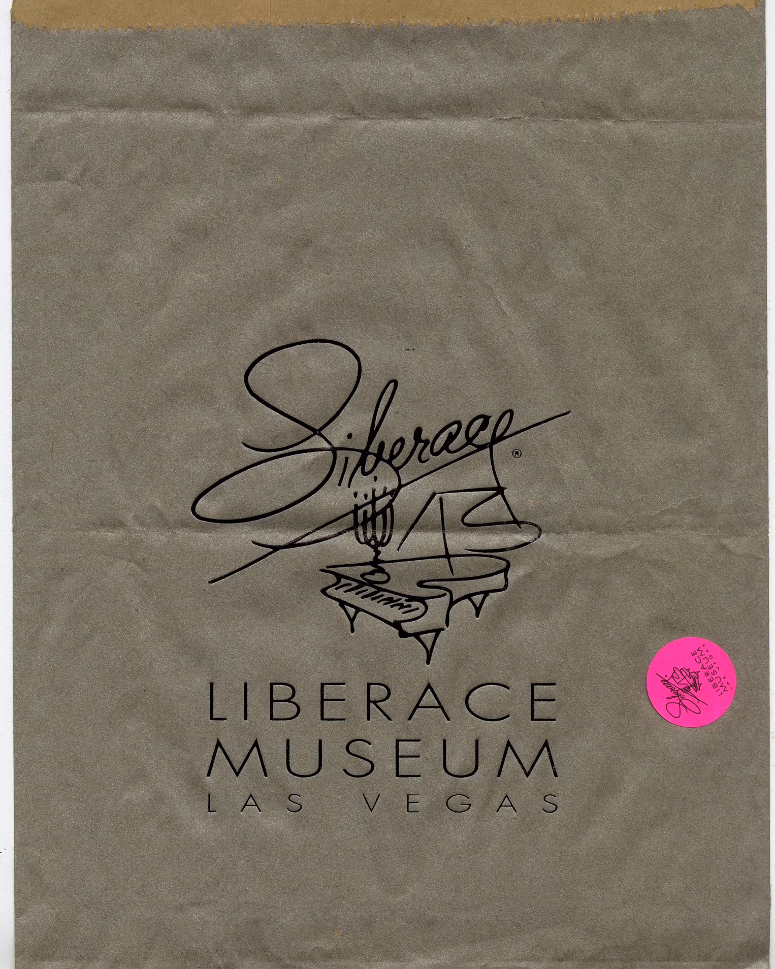 A brown paper back with the logo of the Liberace Museum Las Vegas. A fine line illustration of a piano with "Liberace" written in cursive handwriting on top. There is a pink sticker at the corner of the paper bag with the same logo but much smaller. 