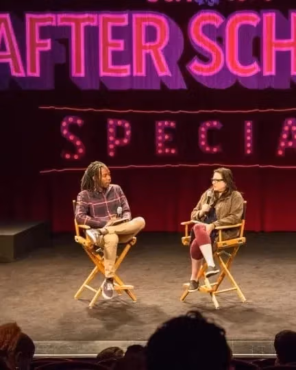 Two people sit on a stage in directors chairs in front of a curtain with the words "After School Special" in pink behind them. There's a crowd facing the stage in the foreground.