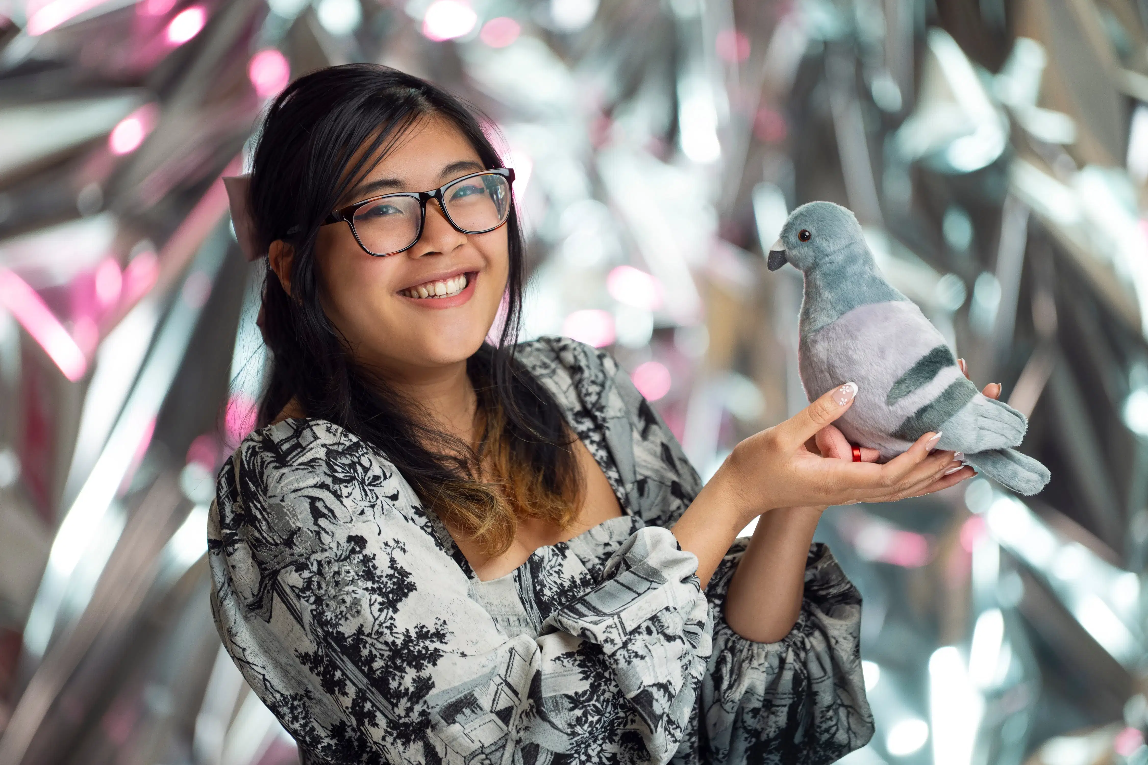 A girl with glasses and a bow in her hair holds a pigeon plus and smiles at the camera.