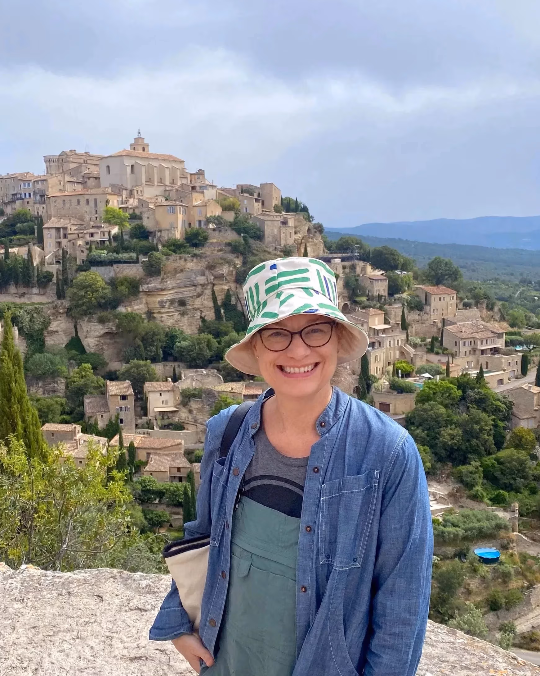 A woman in a white and blue bucket hat and a blue shirt, smiles and stands in front of