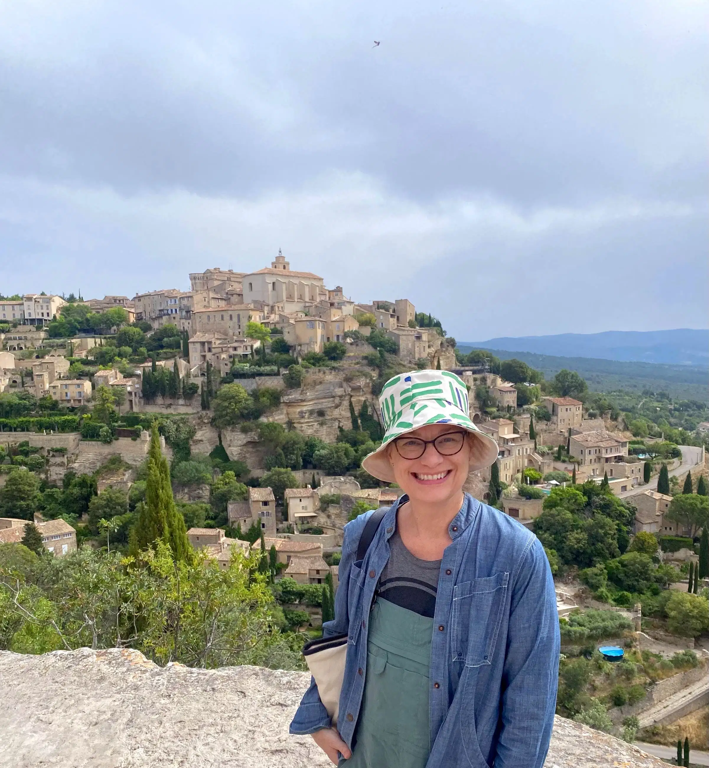 A woman in a white and blue bucket hat and a blue shirt, smiles and stands in front of