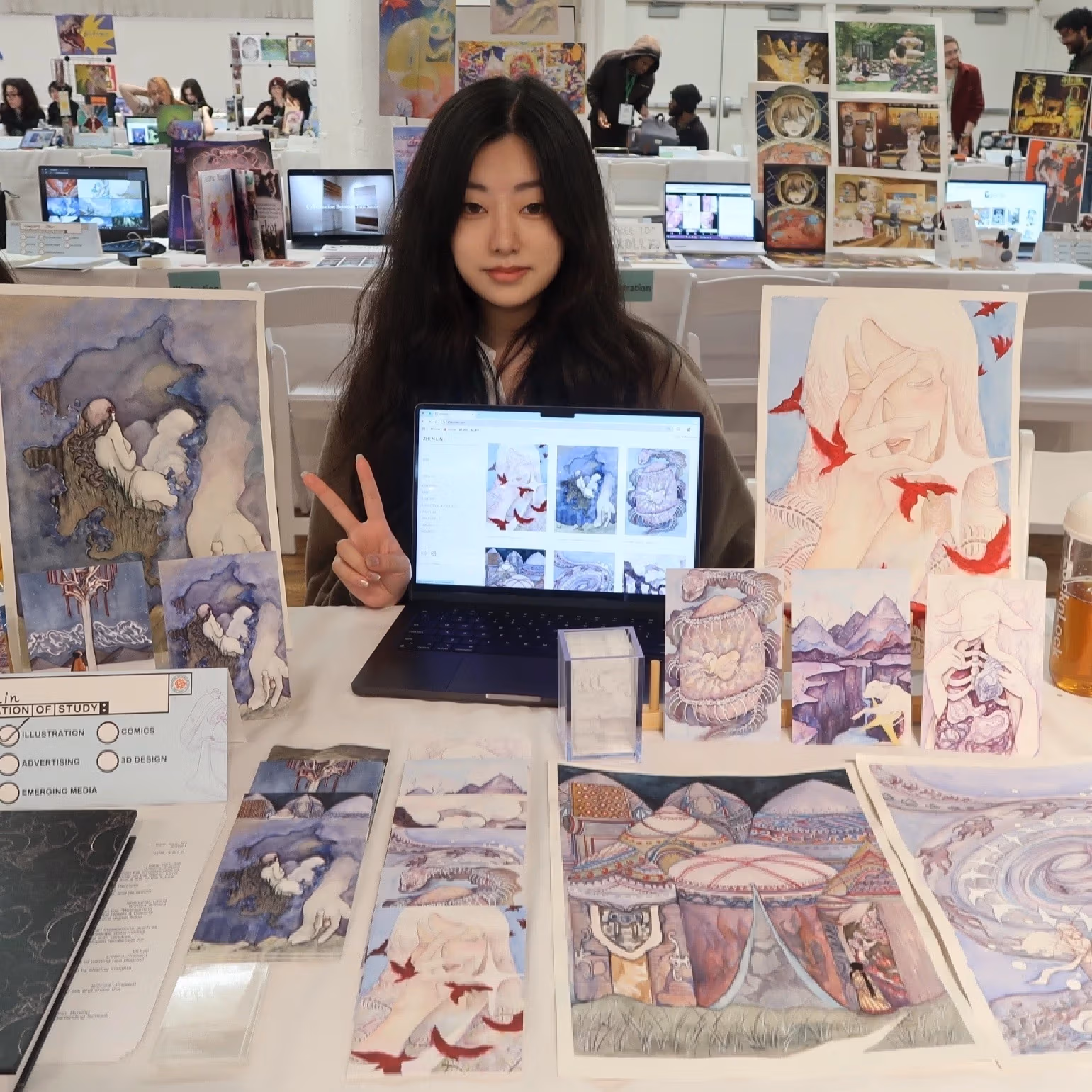 A girl poses at a table displaying a variety of artwork.