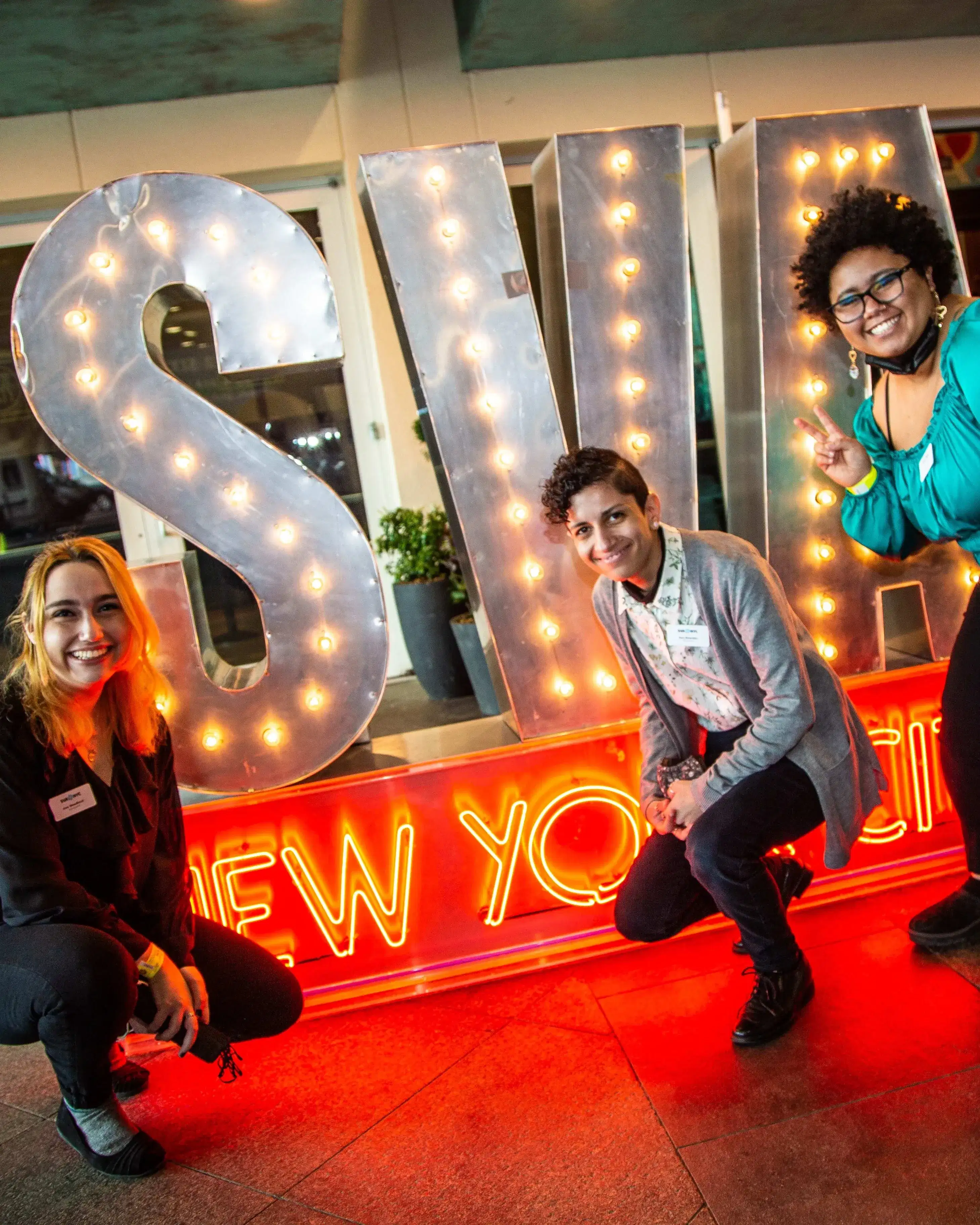 Three people smile and pose in front of a neon SVA New York City sign