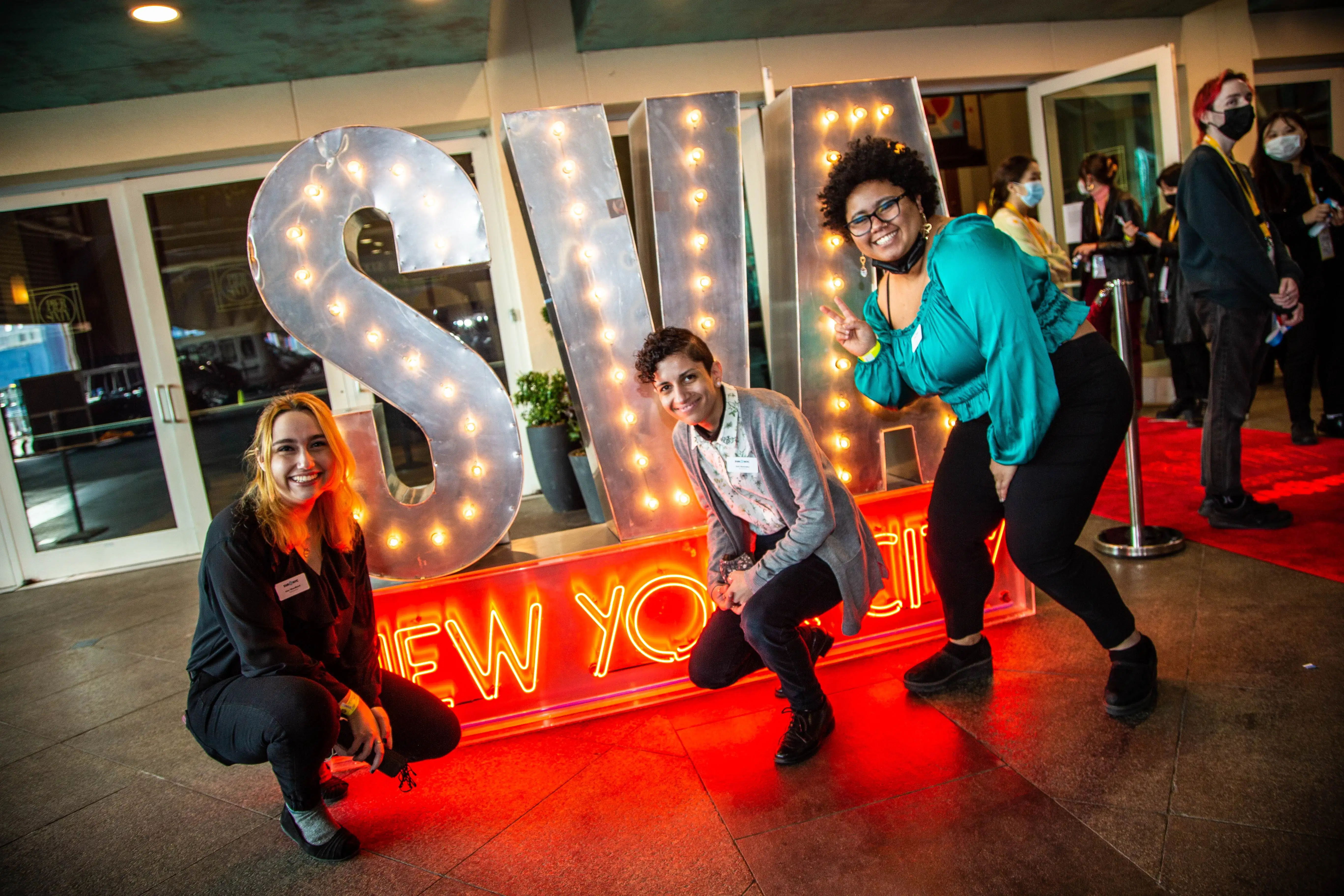 Three people smile and pose in front of a neon SVA New York City sign