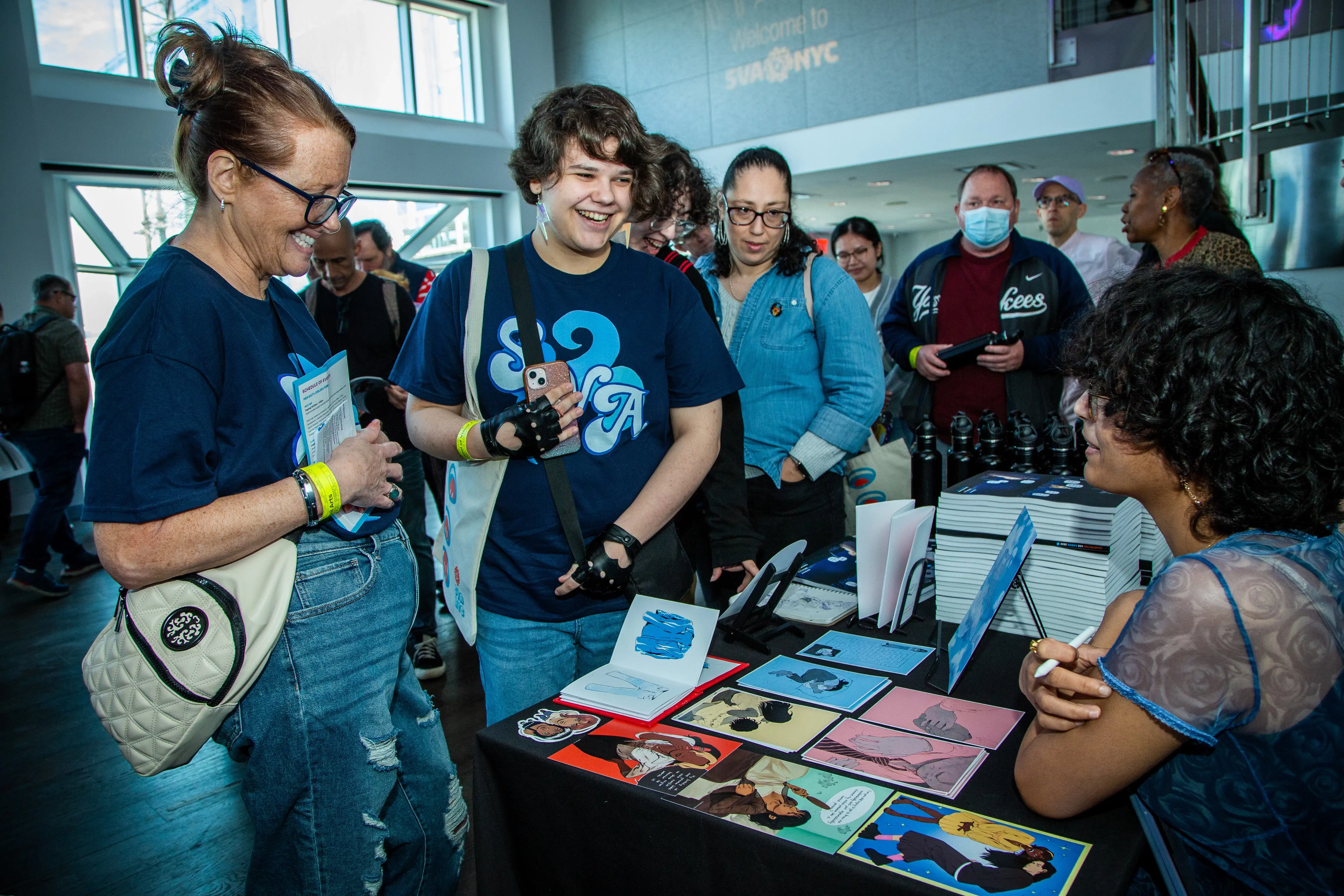 Two people wearing matching shirts are smiling while standing in front of a table of colorful art illustrations