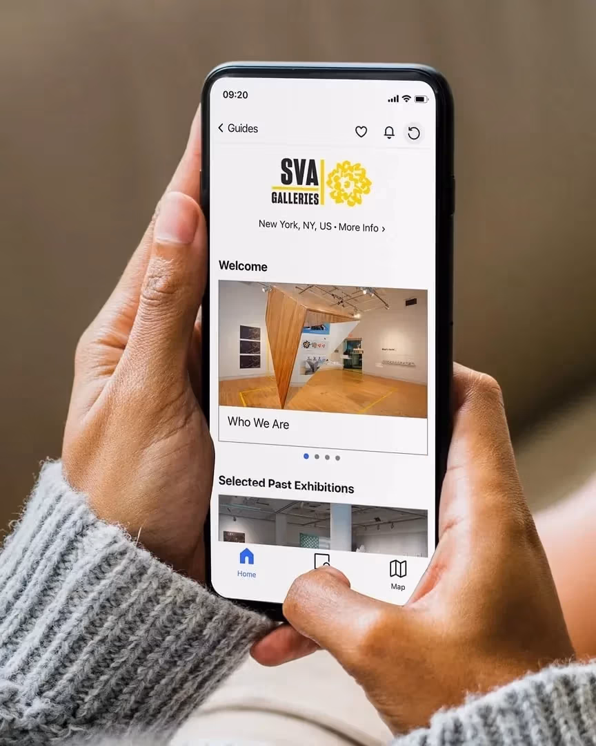 Over-the-shoulder photograph of a person holding a phone in their hand, displaying the SVA Galleries Digital Gallery Guide on the Bloomberg Connects platform.