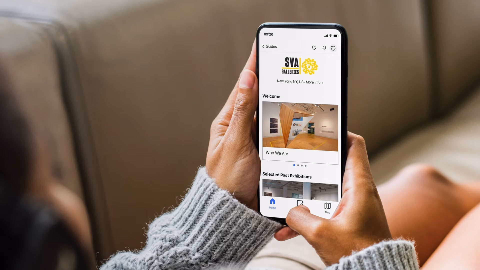 Over-the-shoulder photograph of a person holding a phone in their hand, displaying the SVA Galleries Digital Gallery Guide on the Bloomberg Connects platform.