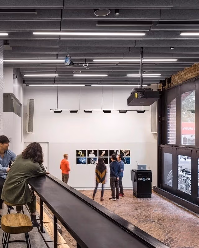 A large room with high ceilings and windows that go from floor to ceiling on the right side of the space. The photo is taken from a mezzanine looking down on the large room. Photographic prints hang on the central wall, with several people gathered around them in the distance. In the foreground, 2 people stand on the mezzanine looking at the same prints. The room has white walls with a dark ceiling and track lighting illuininating the wall of prints. A black podium with a computer atop it sits to the right of th e prints with the SVA NYC logo in blue on the front of it.  
