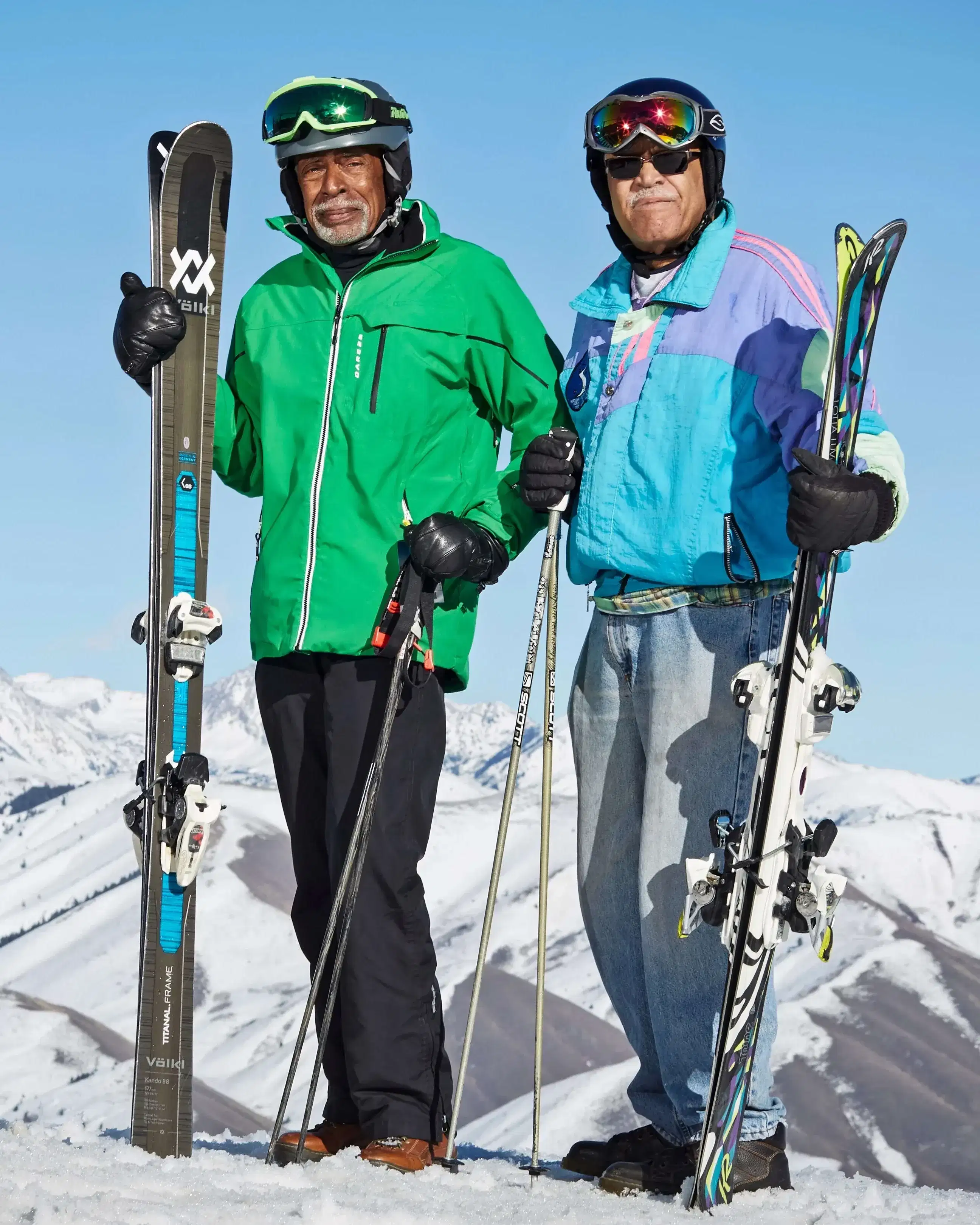 A color photo of two male skiers on the top of a snow-covered mountain. They both have dark skins, are wearing ski outfits, and carrying ski equipment. They are both smiling into the camera.