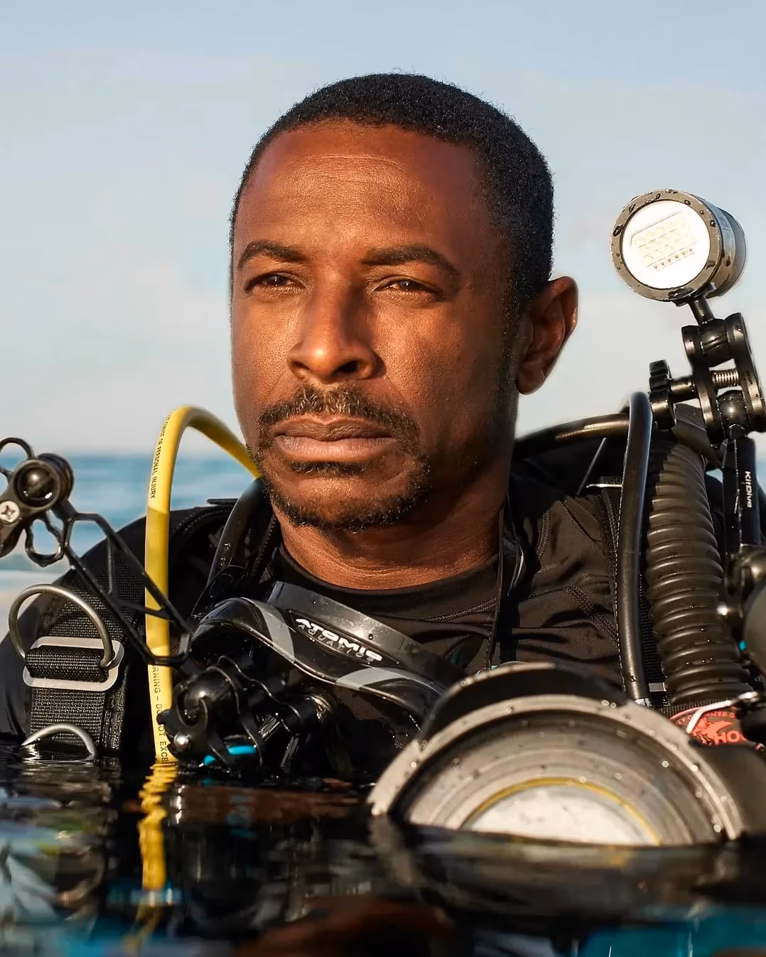 A portrait of a scuba diver in water. He has darker skin and short hair, is wearing a black top, and is equipped with numerous lighting and camera equipments strapped to his body. His body is submerged in water from the chest down.