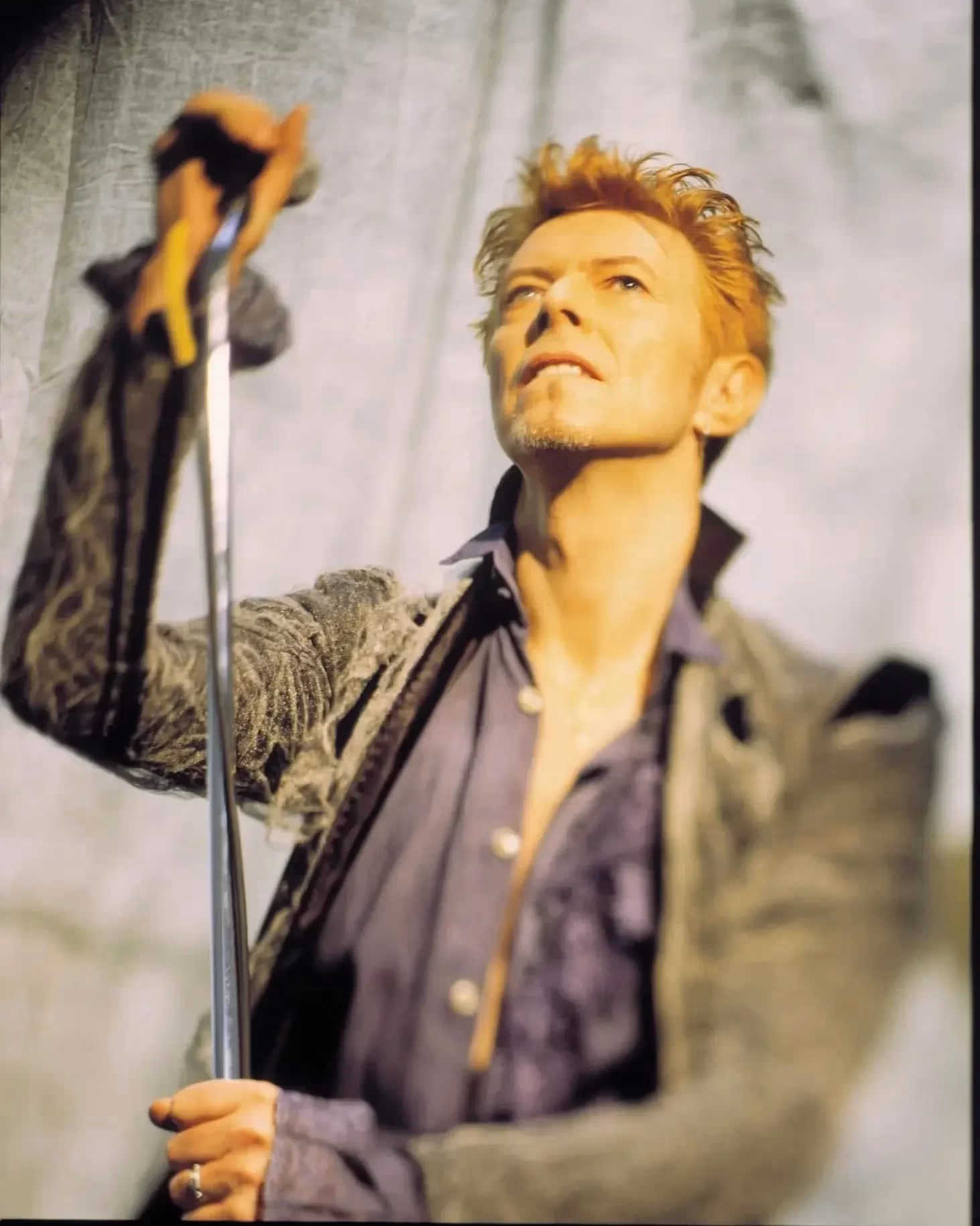 A person with styled light hair and a goatee wears a velvet jacket and shirt, looking upward while holding a microphone stand against a light background.
