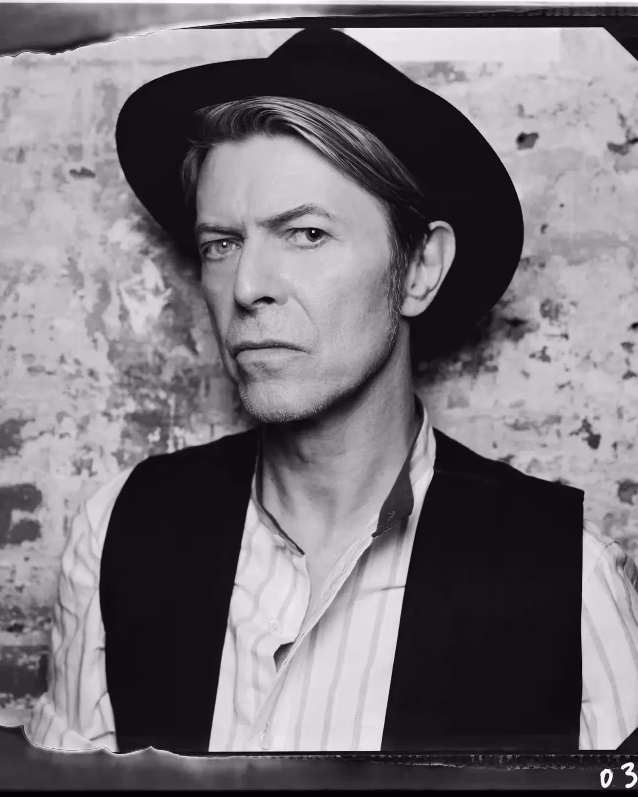 Black and white portrait of a person wearing a brimmed hat and vest, standing against a textured wall. They have a serious, contemplative expression.